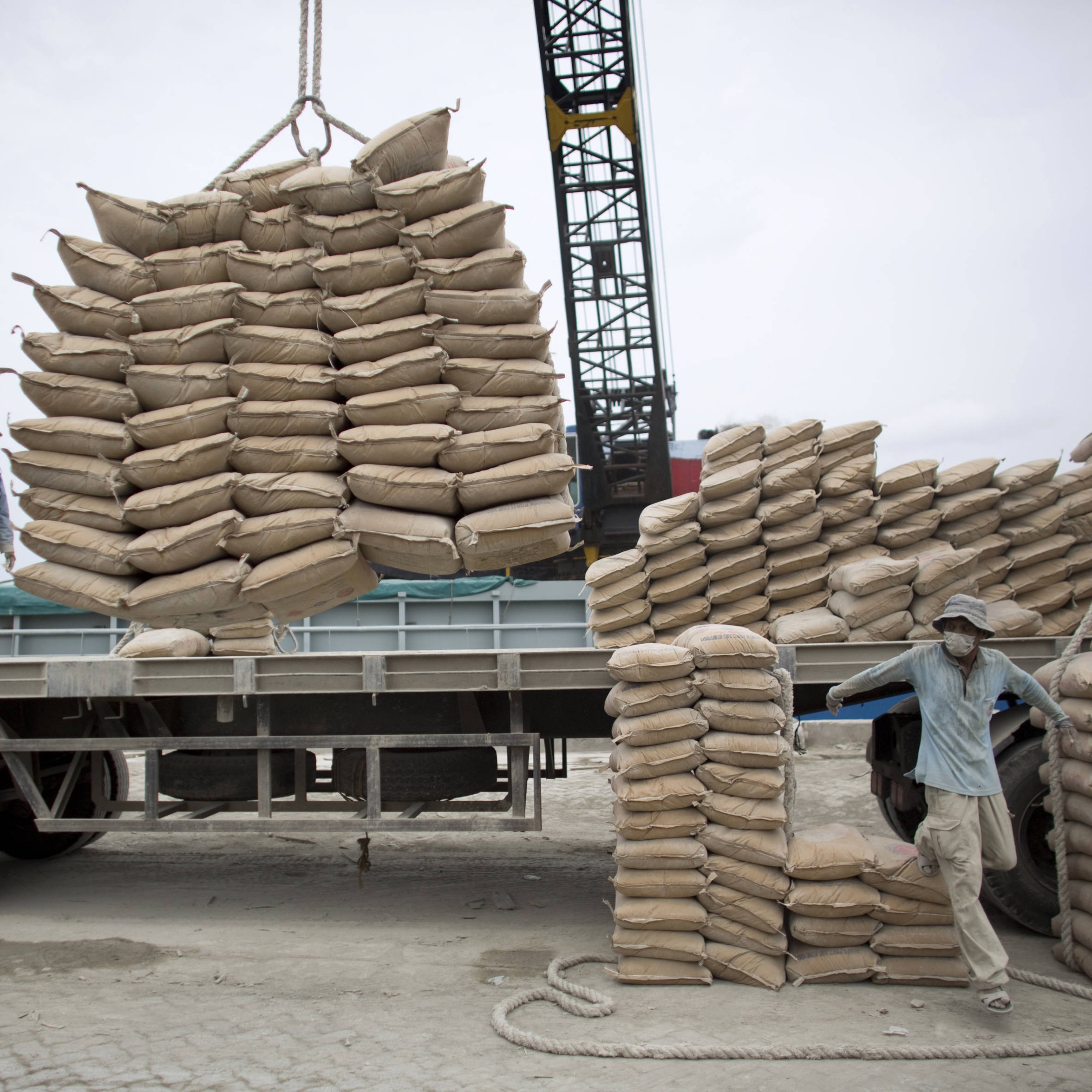 L'image montre une scène de travail au port ou dans un chantier. On y voit des hommes qui manipulent des sacs de sable ou de ciment. Certains sont en train de charger des sacs sur un camion à l'aide d'une grue, tandis que d'autres semblent organiser les sacs déjà empilés. L'environnement est industriel, avec des matériaux de construction et des équipements lourds visibles en arrière-plan. Le ciel est nuageux, ce qui donne une ambiance un peu grise à la scène.