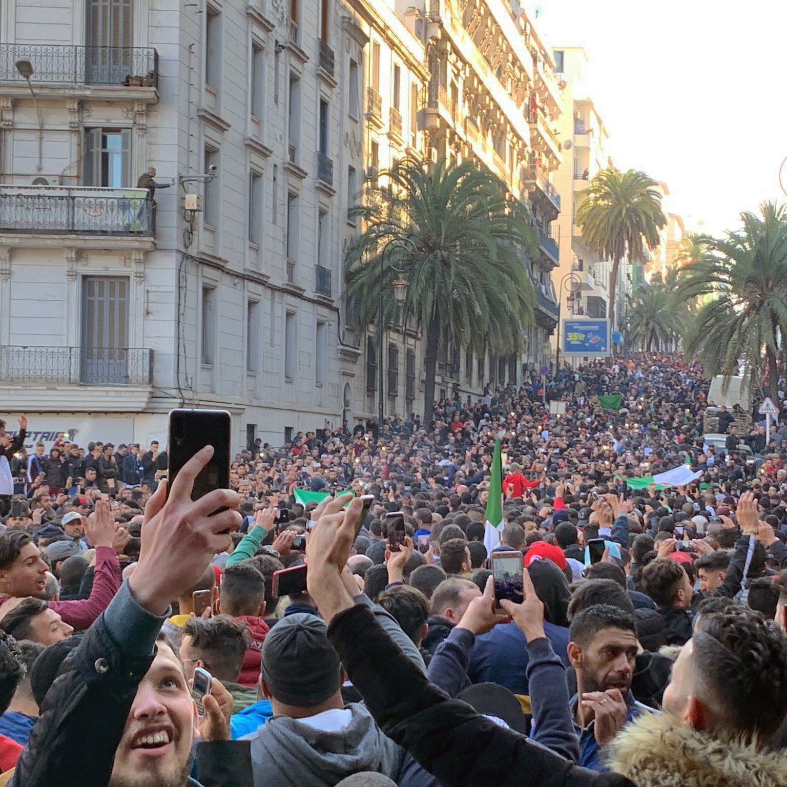 L'image montre une grande foule de personnes rassemblées dans une rue. Les participants semblent manifester, brandissant des drapeaux et levant des mains, certains tenant des téléphones pour filmer. On peut voir des bâtiments en arrière-plan, ainsi que des palmiers qui ajoutent une touche estivale à la scène. L'atmosphère semble dynamique et engagée, illustrant un moment de solidarité ou de protestation.