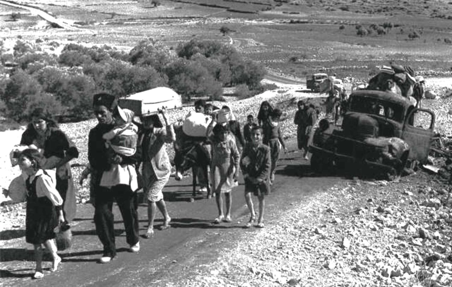 L'image montre un groupe de personnes marchant le long d'une route, probablement en situation de déplacement ou d'exode. On peut voir des adultes portant des enfants et des charges, tandis que des enfants marchent également. À l'arrière-plan, des véhicules abandonnés et un paysage aride s'étendent, suggérant une situation difficile. L'atmosphère semble grave et chargée d'émotion, témoignant des épreuves que traversent ces personnes.
