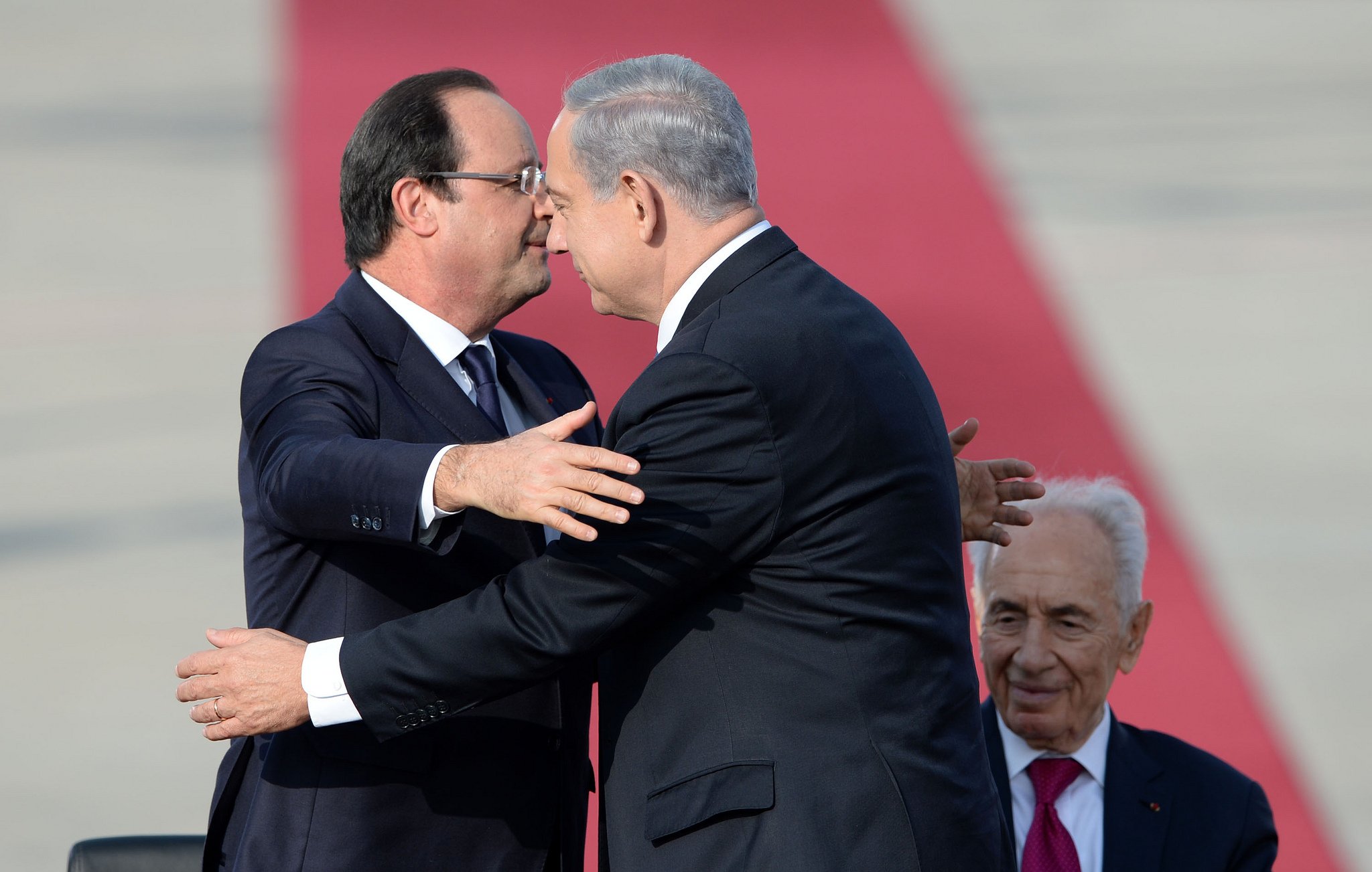 L'image montre deux hommes qui se serrent dans les bras dans un geste amical, tandis qu'un troisième homme est assis derrière eux, regardant la scène. Les deux hommes à l'avant portent des costumes, et on peut notamment voir l'un d'eux pencher la tête en signe de proximité. L'arrière-plan est basé sur une surface rouge, suggérant un événement officiel ou cérémoniel. L'atmosphère semble conviviale et respectueuse.