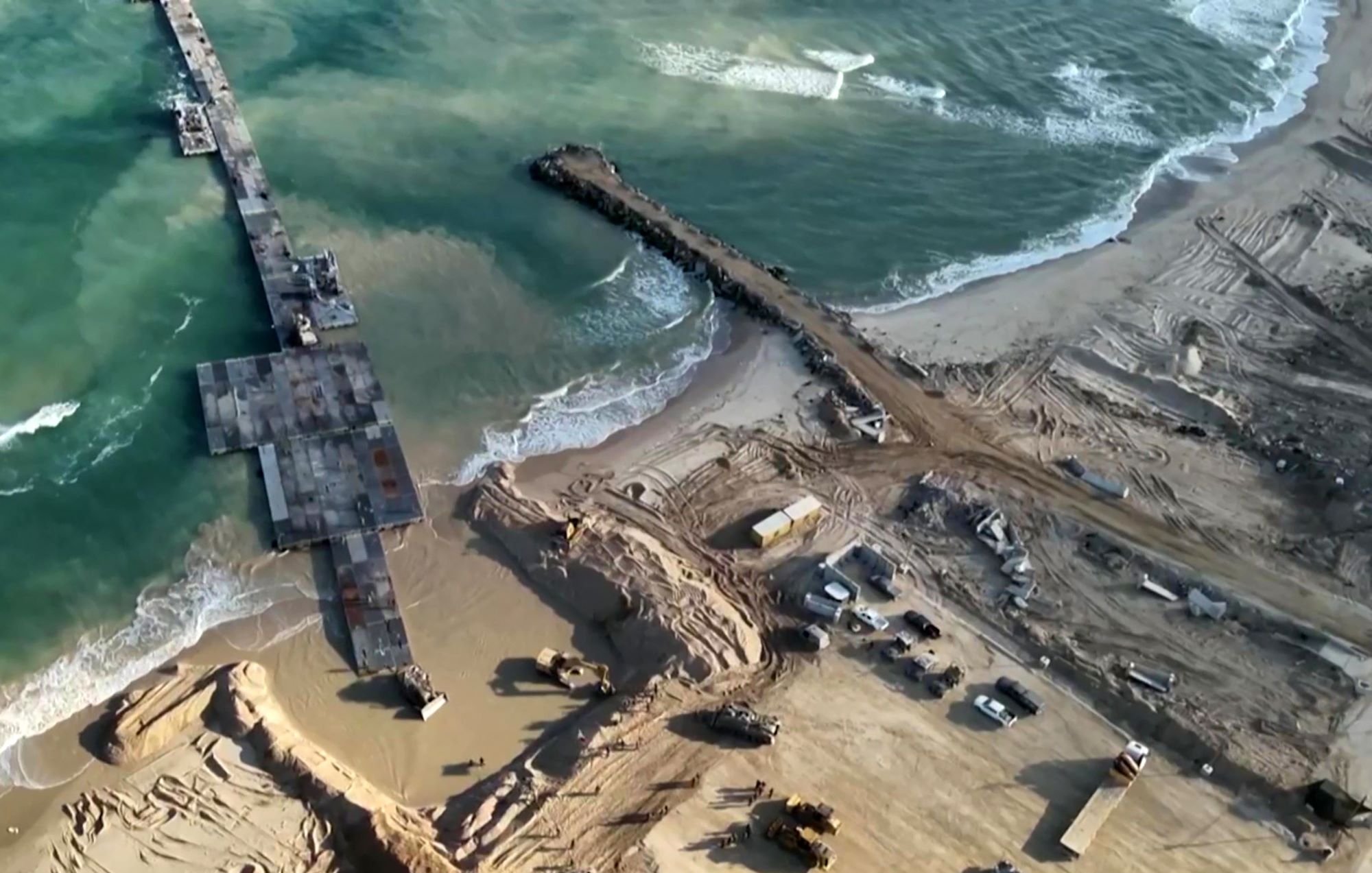 L'immagine mostra una vista aerea di una zona costiera in fase di costruzione o ristrutturazione. Si possono vedere macchine da lavoro e attrezzature presenti sulla spiaggia e nelle vicinanze, mentre un molo si estende verso il mare. Le onde del mare si infrangono sulla riva, e il terreno circostante appare movimentato e scavato, con evidenti segni di lavori in corso. La luce naturale esalta i dettagli del paesaggio, creando un contrasto tra l'acqua blu e la sabbia.