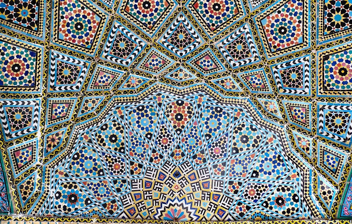 L'image montre un magnifique plafond orné de motifs géométriques et floraux colorés. Les couleurs vives, comme le bleu, le rouge, le jaune et le noir, sont disposées de manière symétrique, créant un effet visuel captivant. Les motifs complexes, typiques de l'architecture islamique, illustrent un savoir-faire artisanal exceptionnel. Ce type de décoration est souvent trouvé dans des monuments historiques et des mosquées.