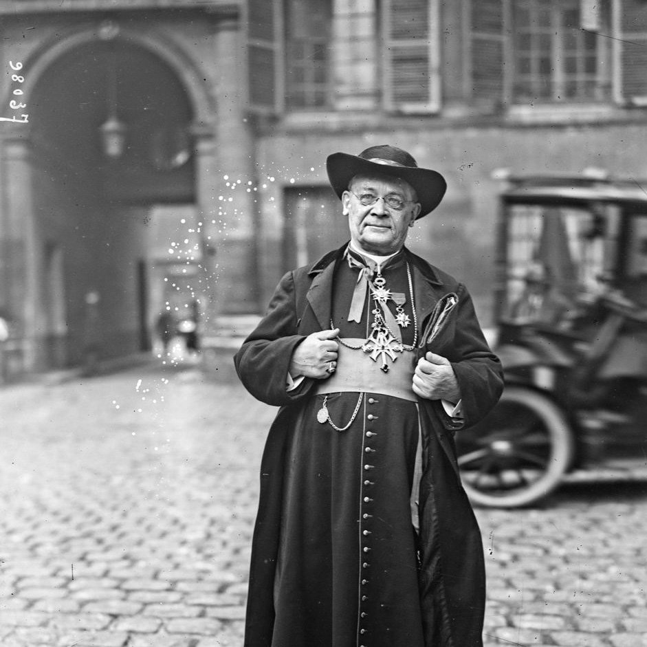 L'image montre un homme en tenue traditionnelle, probablement un ecclésiastique. Il porte une longue robe noire avec des ornements et des boutons sur le devant. Son chapeau est large et il semble poser avec une certaine dignité, les bras légèrement écartés. En arrière-plan, on aperçoit des bâtiments anciens et un véhicule à roues, ce qui suggère une ambiance historique. L'environnement de la rue est pavé, ajoutant à l'atmosphère d'époque.