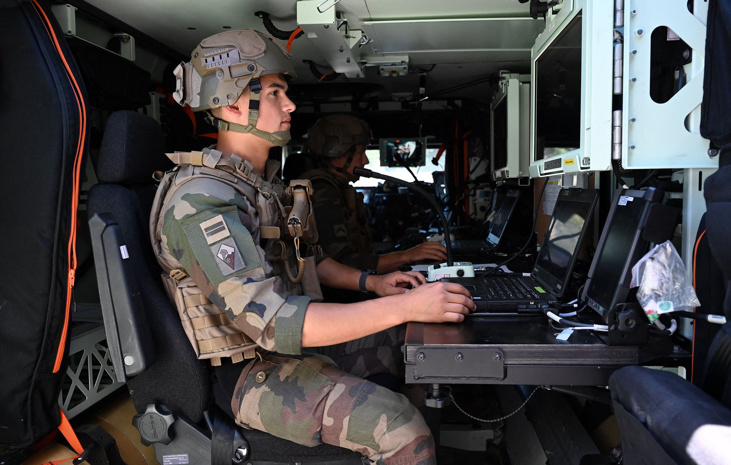 L'image montre deux soldats à l'intérieur d'un véhicule militaire. Ils portent des uniformes camouflés et des casques. L'un des soldats est assis devant un ordinateur portable, concentré sur son travail. L'intérieur du véhicule est équipé de plusieurs écrans et dispositifs technologiques. L'atmosphère semble sérieuse, indiquant une mission ou une opération en cours.