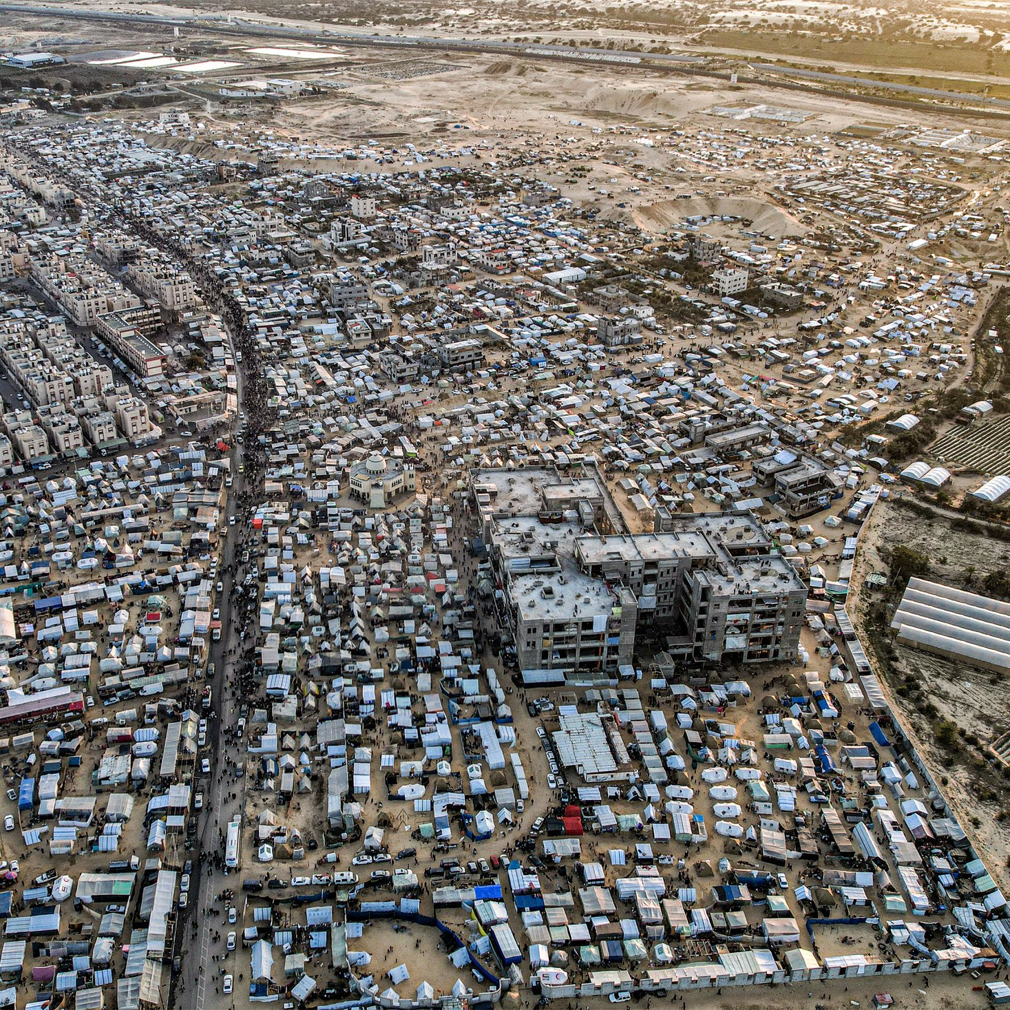 این تصویر نمای هوایی از یک منطقه شلوغ و پرجمعیت است. در اینجا تعداد زیادی ساختمان و خانه های کوچک دیده می‌شود که در کنار هم قرار گرفته‌اند. خیابان‌هایی که به نظر می‌رسد مردم در آن تردد دارند، میانه تصویر را تقسیم کرده‌اند. همچنین، در برخی نواحی، چادرها و سازه‌های موقت دیده می‌شوند که نشان‌دهنده زندگی در شرایط سخت و غیرفرهنگین است. این تصویر، وضعیتی را به نمایش می‌گذارد که بیانگر چالش‌های اجتماعی و اقتصادی است.