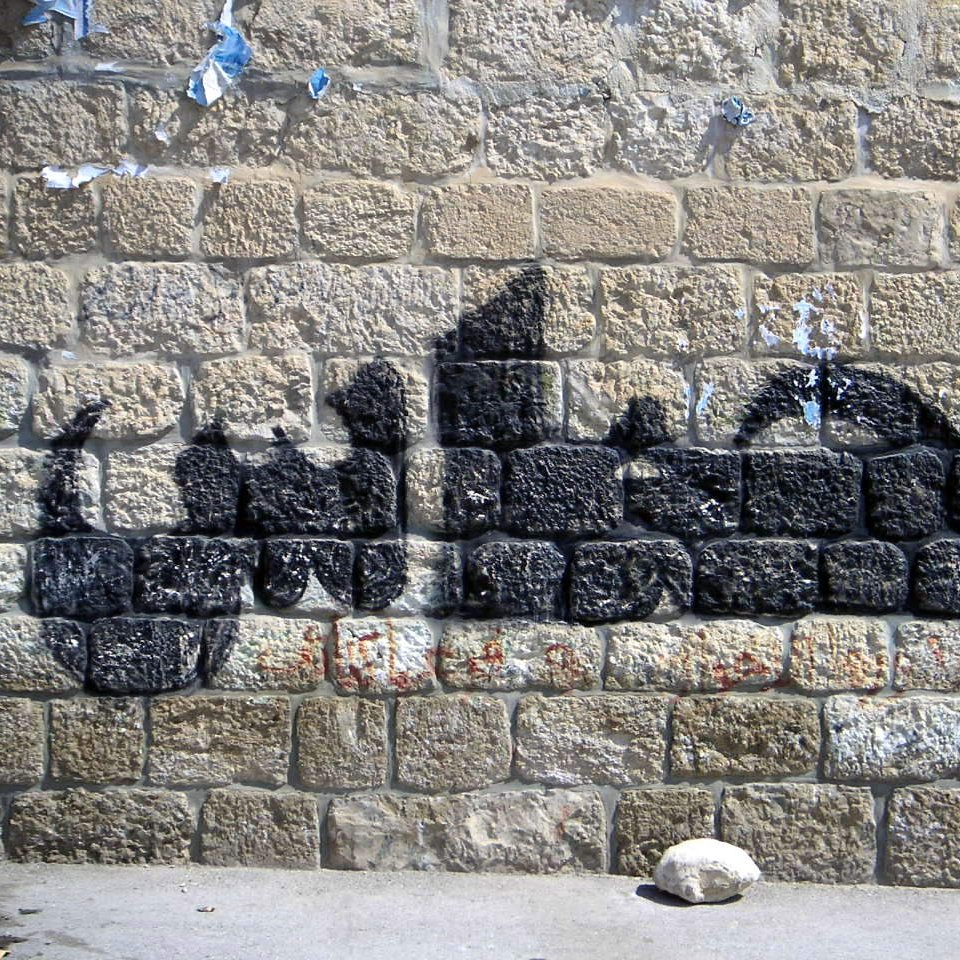 L'image montre un mur de pierres avec un graffiti en forme de silhouette noire. Le mur semble ancien et légèrement abîmé, avec des morceaux de papier déchirés. La silhouette créée par le graffiti évoque des formes humaines, mais sans détails précis. Les pierres du mur sont bien visibles et ajoutent une texture rustique à l'ensemble.