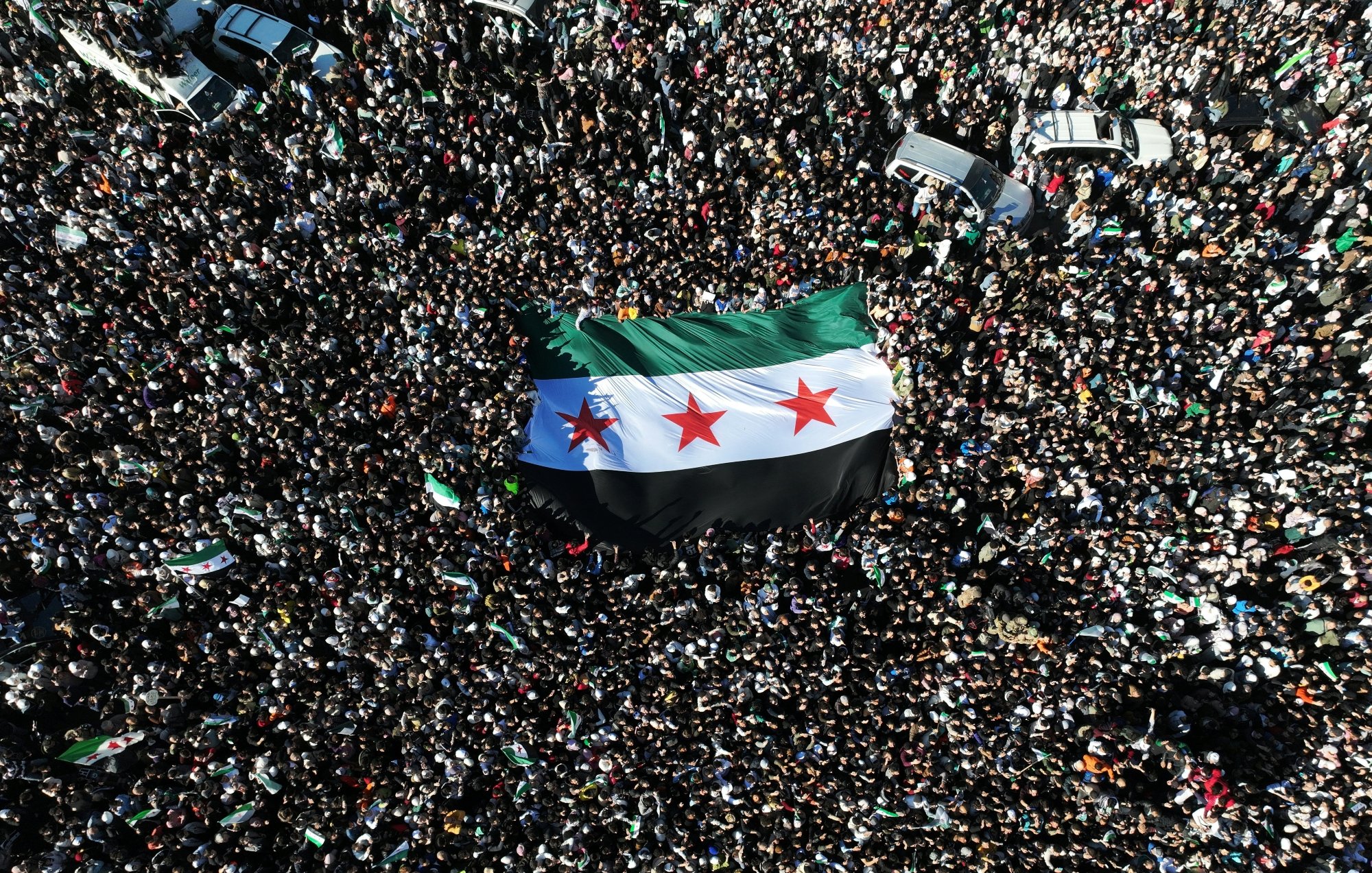 Une foule dense brandit un grand drapeau noir, blanc et vert avec trois étoiles rouges.