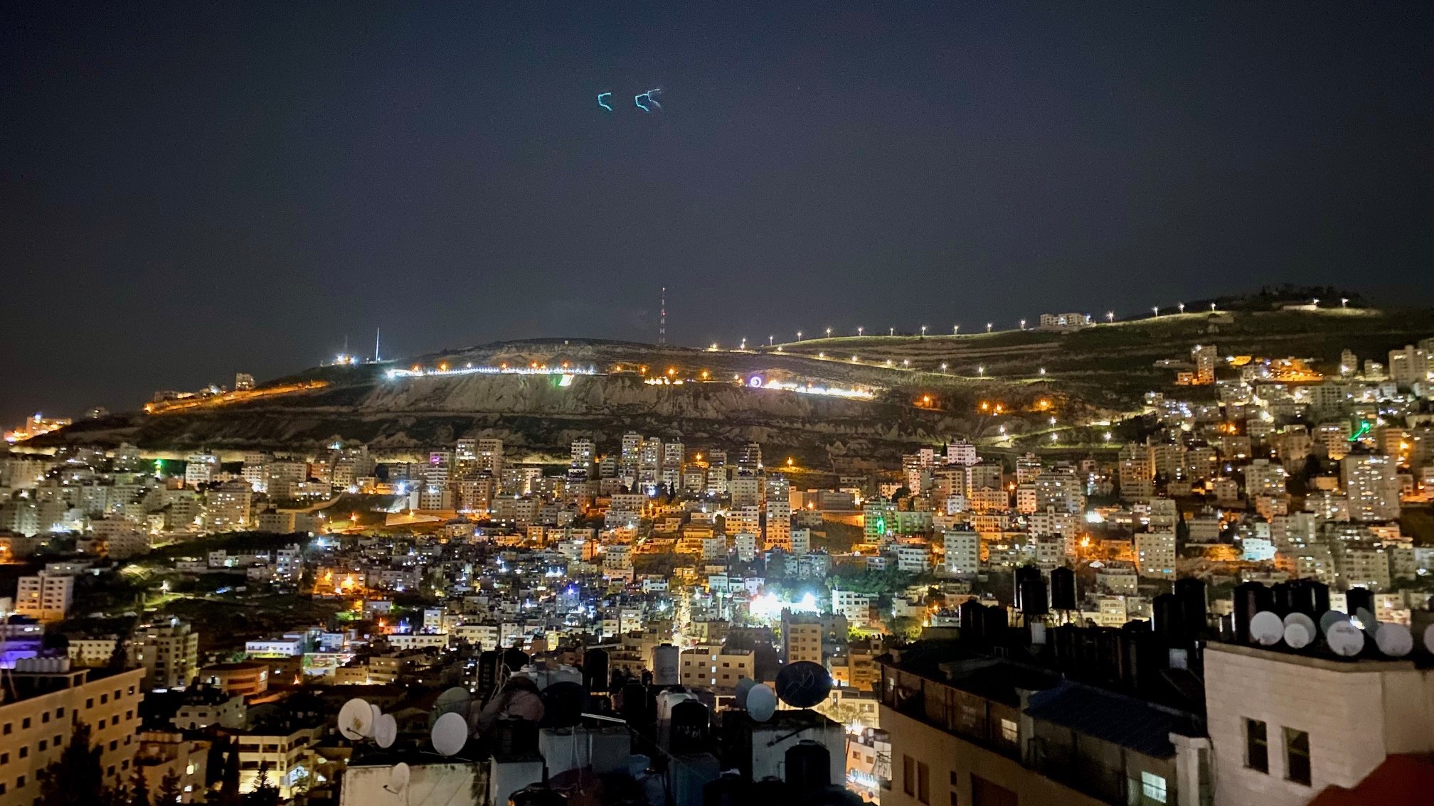 The image shows a nighttime view of a cityscape, likely on the slope of a hill or mountain. The scene is illuminated with numerous lights from buildings, giving it a vibrant and lively atmosphere. In the foreground, there are rooftops with antennas or satellite dishes visible, while the background features the illuminated hillside, possibly with telecommunications towers or other structures visible at the top. The sky is dark, suggesting it's a clear night with some stars or light reflections. Overall, the image conveys a sense of urban life and a stunning nocturnal landscape.