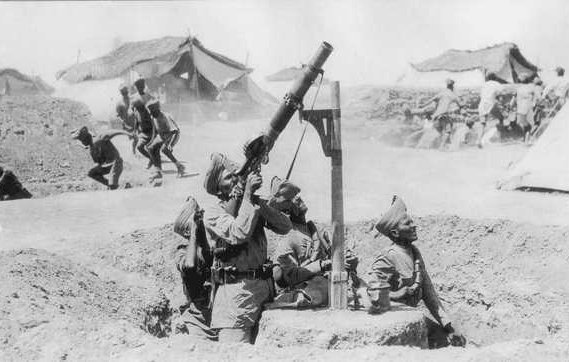 L'image en noir et blanc montre un groupe de soldats en position de tir. Un soldat est en train d'opérer un mortier, levé vers le ciel, tandis que deux autres soldats se trouvent à proximité, probablement prêts à aider ou à surveiller la situation. En arrière-plan, on peut voir des tentes militaires, indiquant qu'ils sont dans un camp. D'autres soldats semblent en mouvement, suggérant une activité intense ou une opération en cours. L'atmosphère générale de l'image évoque un contexte militaire et historique.