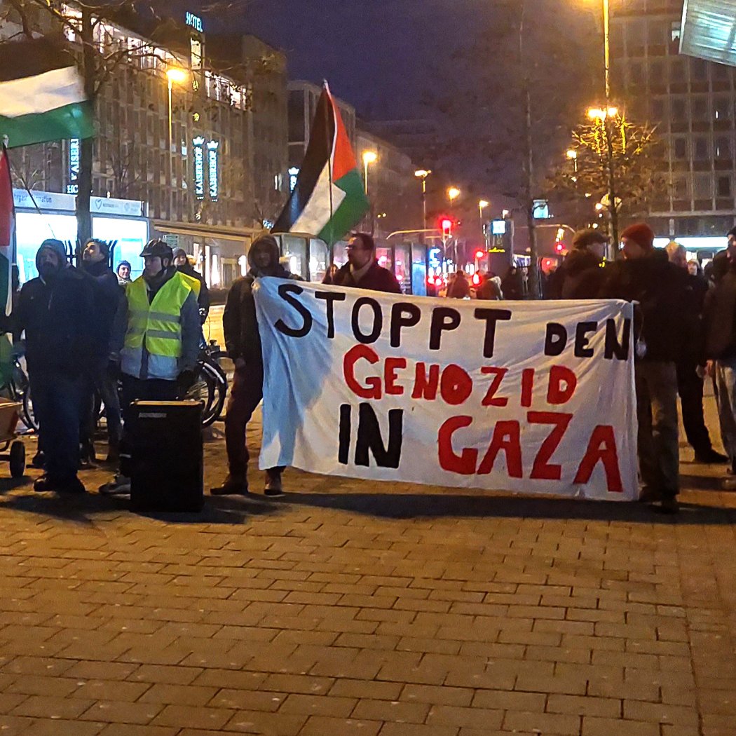 L'image montre un groupe de manifestants rassemblés dans une rue, tenant une grande banderole sur laquelle il est écrit "STOPPT DEN GENOZID IN GAZA". Ils portent des drapeaux palestiniens et semblent exprimer leur soutien à Gaza. L'atmosphère est sérieuse, et il est probable que la manifestation s'inscrive dans un contexte de protestation contre des violences ou des injustices en Palestine. Les lumières des bâtiments environnants suggèrent qu'il s'agit d'une manifestation en soirée.