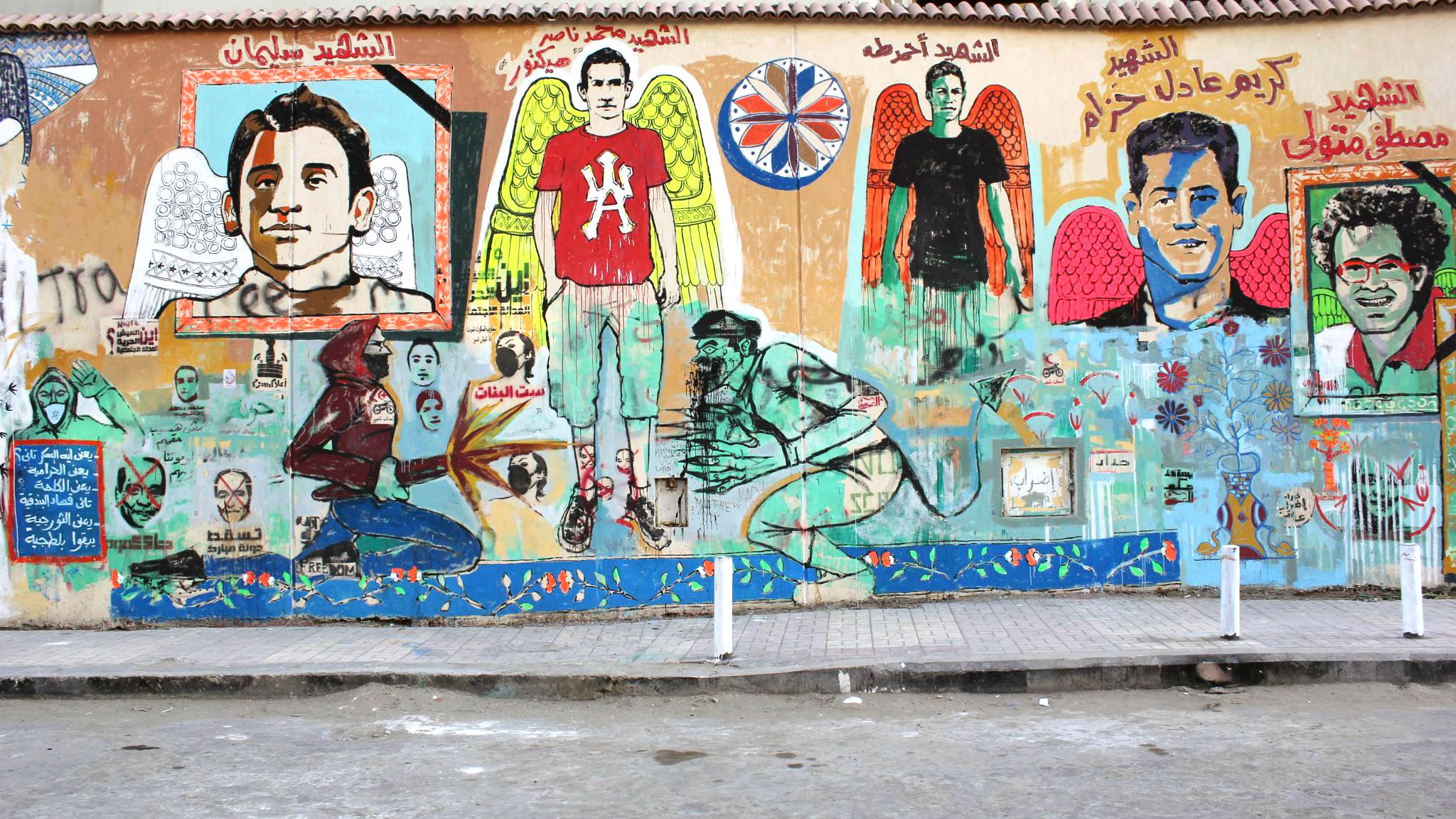 L'image représente une fresque murale colorée sur un mur. On y voit plusieurs portraits d'individus, accompagnés de motifs d'ailes et de détails symboliques. Les personnages ont des expressions variées, et des écritures en arabe sont visibles en haut de la fresque. Le fond présente des éléments artistiques tels que des fleurs et d'autres motifs graphiques. L'ambiance générale est vibrante et évoque un message fort, probablement en lien avec la culture locale ou un hommage à des figures importantes. Le sol devant le mur est en béton, et on peut y voir des supports en métal.