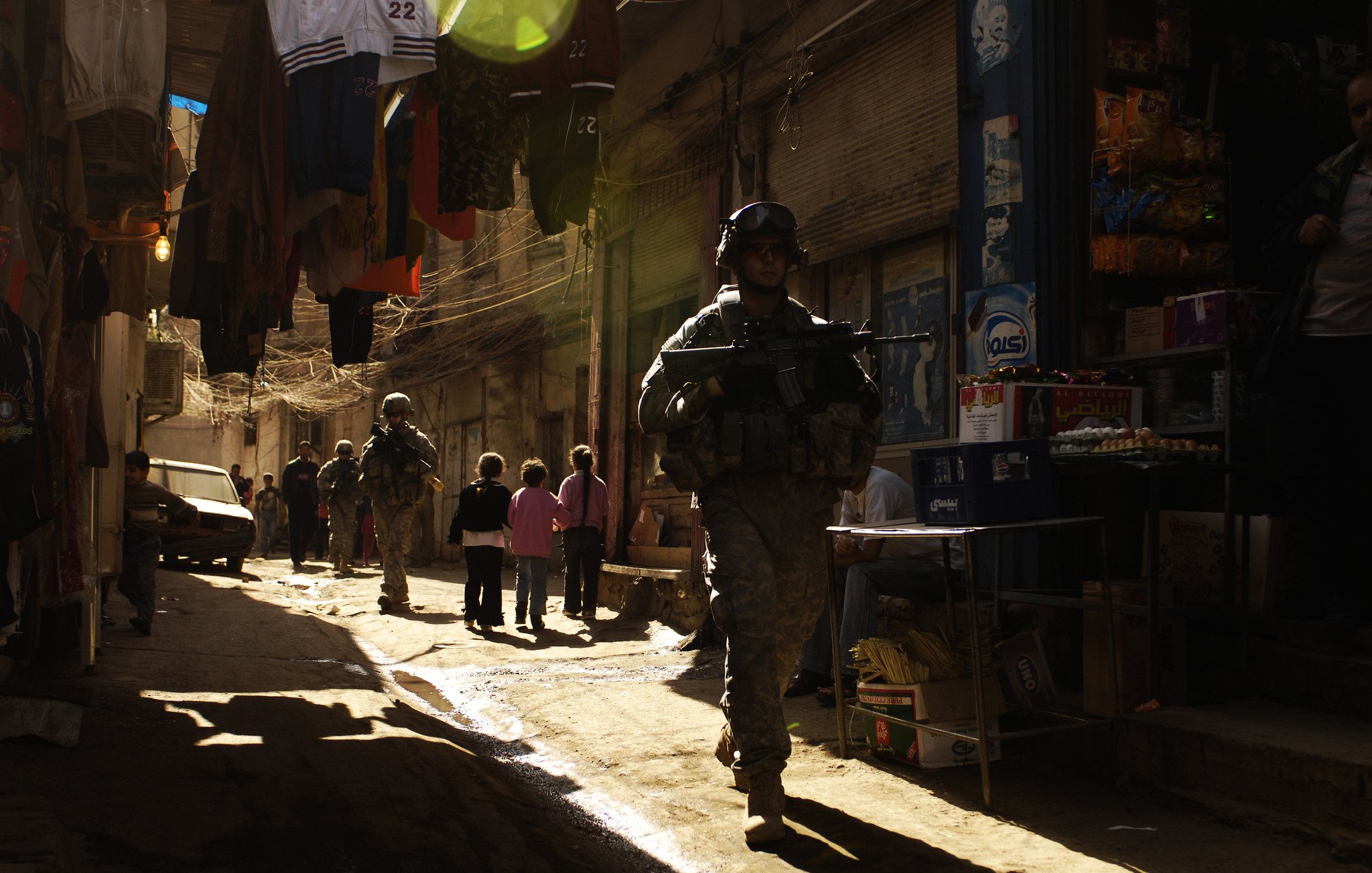 La imagen muestra a soldados patrullando en un callejón estrecho y iluminado por la luz del sol. A los lados, hay ropa colgada y tiendas de barrio que parecen ofrecer diversos productos. Un grupo de niños está presente en la escena, observando a los soldados. La atmósfera es tensa, dado el contexto militar, pero la luz crea un efecto visual interesante en el entorno.