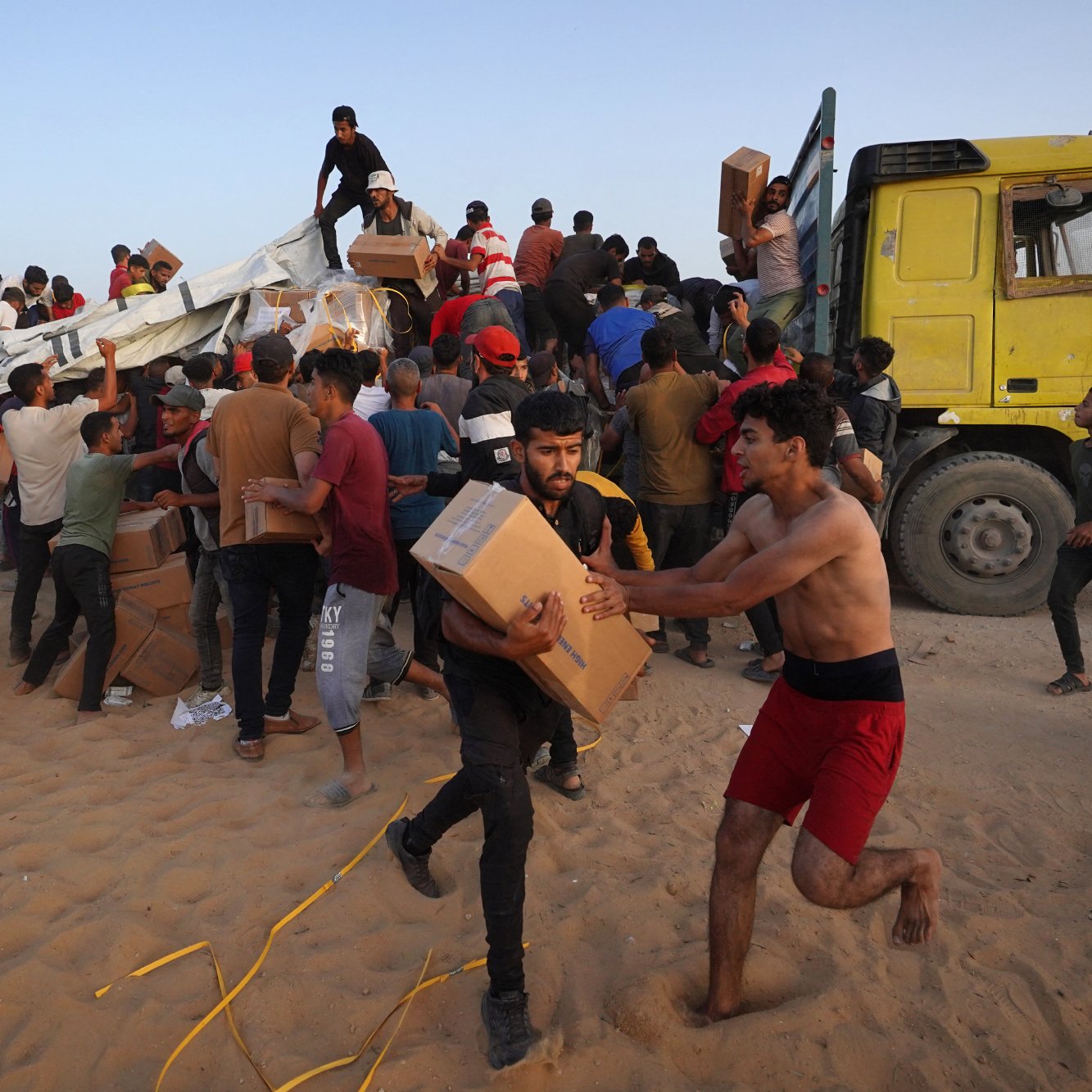 L'immagine mostra un gran numero di persone, principalmente giovani uomini, che sono radunati attorno a un camion giallo. Alcuni di loro sembrano scaricare scatole, mentre altri stanno trasportando i pacchi. L'ambiente appare polveroso e le persone sono visibilmente attive e impegnate nell'operazione di distribuzione, con espressioni di urgenza. Alcuni individui sono a torso nudo e indossano abiti vari. Si percepisce un'atmosfera di collaborazione nel tentativo di ricevere e distribuire i rifornimenti.