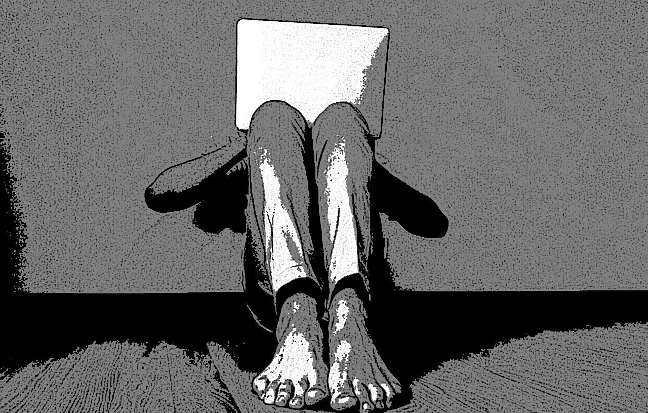 L'image montre une personne assise par terre, adossée à un mur. Elle tient un ordinateur portable devant elle, ce qui cache son visage. Ses jambes sont repliées et ses pieds nus sont visibles. L'ambiance est plutôt sombre et minimaliste, avec un contraste marqué entre les ombres et la lumière.