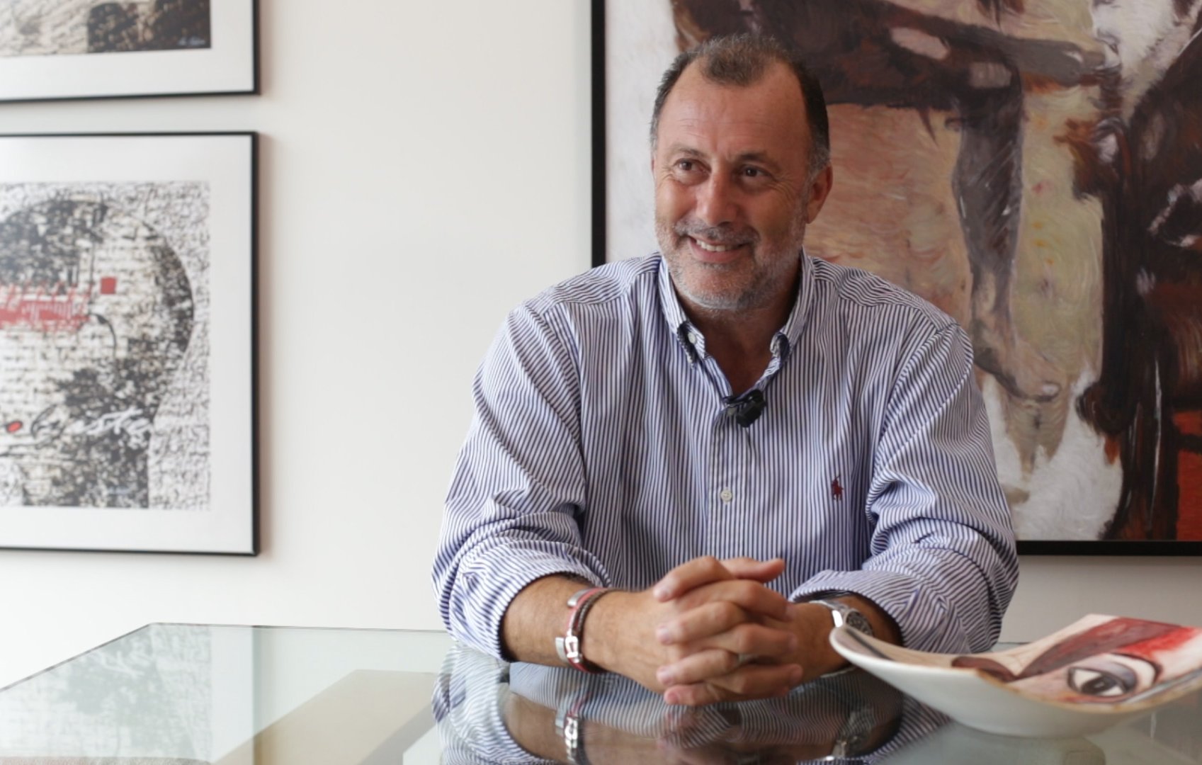 الصورة تظهر رجلًا يجلس على طاولة زجاجية في غرفة ذات ديكور عصري. يبتسم الرجل ويبدو مرتاحًا، بينما خلفه توجد لوحتان فنيتان على الجدار. يعكس الضوء من النوافذ المحيطة الجو العام للاسترخاء والتفاؤل. يبدو أن الرجل يرتدي قميصًا مخططًا بألوان فاتحة، مما يضيف لمسة من الأناقة.