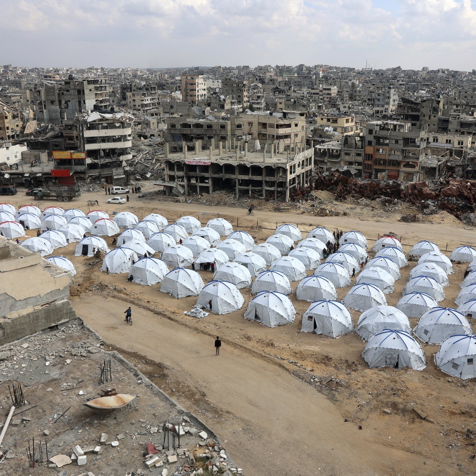 L'immagine mostra un'ampia area con numerosi tende bianche disposte in file ordinate, che rappresentano un campo per sfollati. Sullo sfondo si possono vedere edifici distrutti e in rovina, segno di conflitti o disastri. Il cielo è nuvoloso, contribuendo a un'atmosfera desolante. La scena evidenzia il contrasto tra le tende temporanee, che offrono riparo, e l'ambiente urbano devastato circostante.
