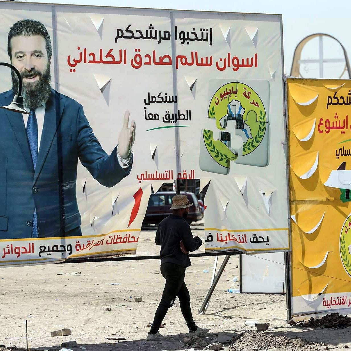 L'image montre un homme marchant devant de grandes affiches électorales dans un environnement urbain. Les affiches présentent des candidats politiques avec leurs noms écrits en arabe, ainsi que des logos et des slogans associés à leur campagne. L'arrière-plan semble désordonné, avec des structures inachevées ou des éléments typiques d'une ville. L'homme semble porter une charge sur sa tête, ajoutant une dimension de vie quotidienne à la scène.