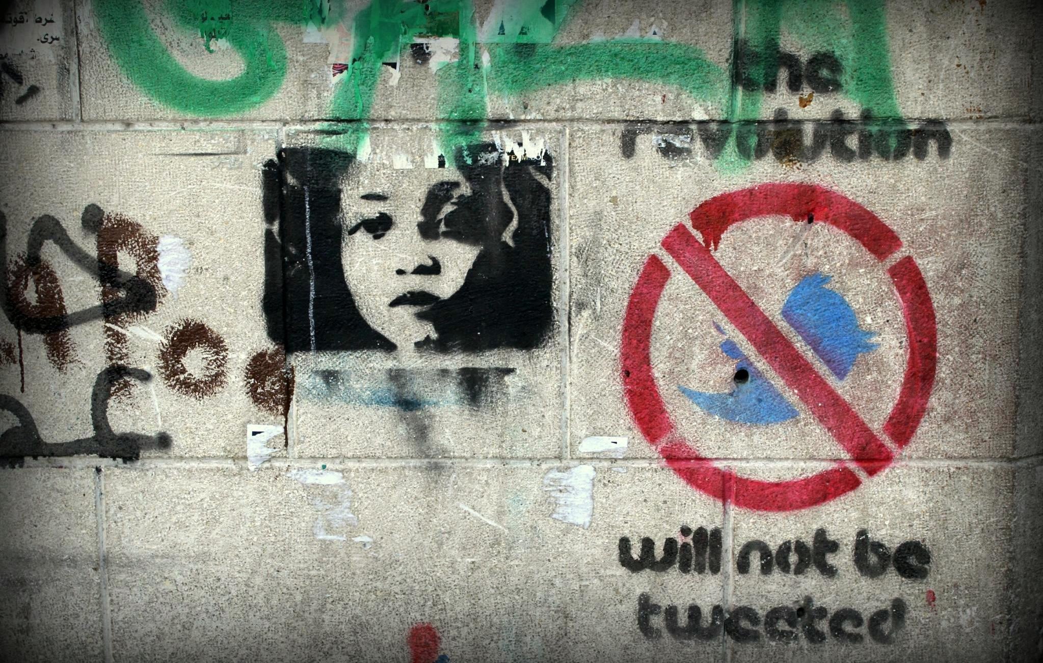 L'image présente un mur couvert de graffitis. On y voit un portrait en noir et blanc d'un enfant qui regarde vers l'observateur. À côté de ce portrait, il y a des inscriptions en couleur, dont l'une mentionne "the revolution" (la révolution) et une autre un symbole barré de l'oiseau bleu, accompagnée du texte "will not be tweeted" (ne sera pas tweeté). L'ensemble évoque des thèmes de révolte et de critique des réseaux sociaux.