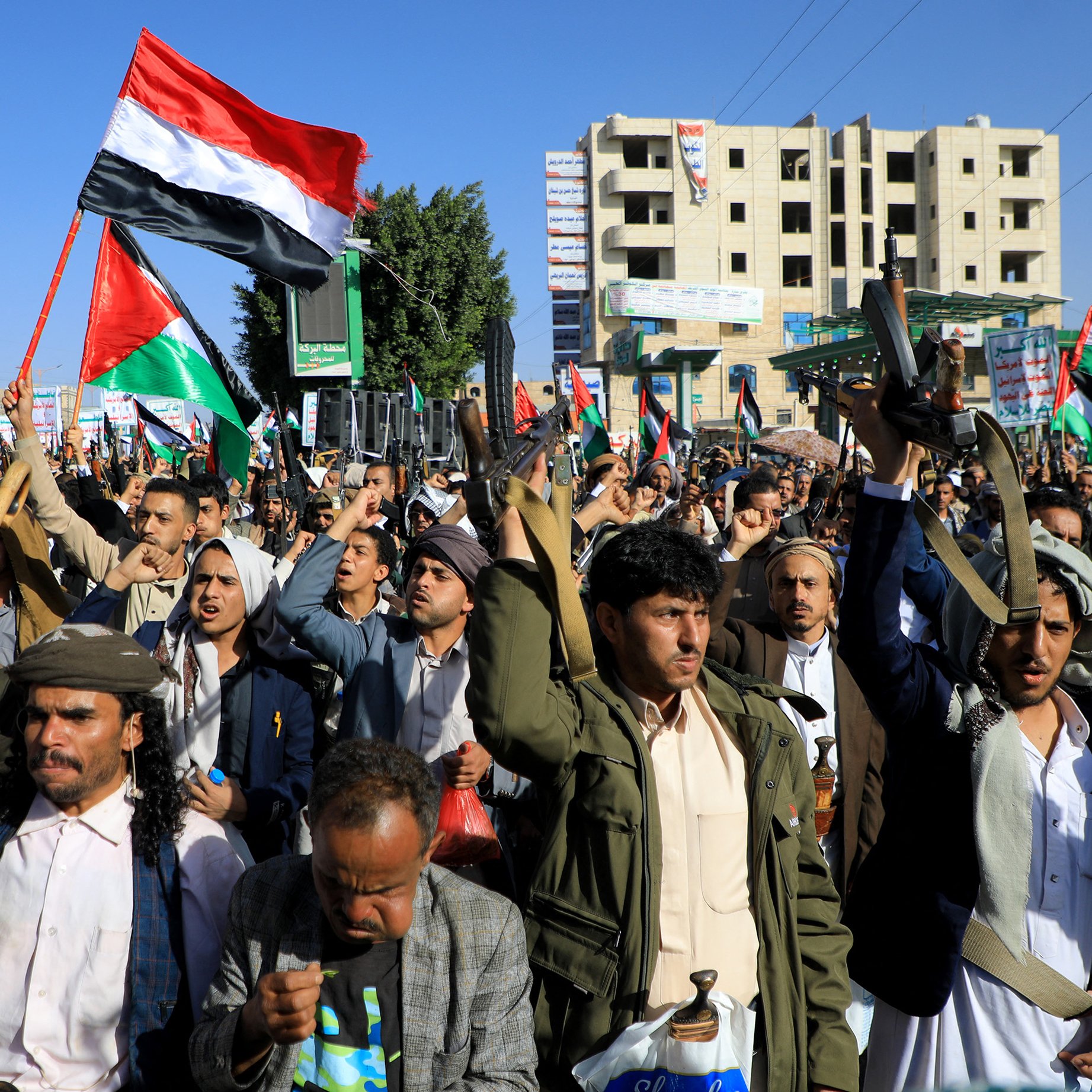 در این تصویر، تعداد زیادی از مردم در خیابانی تجمع کرده‌اند و پرچم‌هایی را در دست دارند. هر فردی ظاهری متعهد و با انرژی به نظر می‌رسد و برخی از آنها در حال فریاد زدن یا شعار دادن هستند. لباس‌ها معمولاً سنتی هستند و برخی از شرکت‌کنندگان در حال بیان نظرات یا احساسات خود در مورد موضوعی خاص به نظر می‌رسند. محیط اطراف شامل ساختمان‌های بلند و تابلوهای تبلیغاتی است که نشان‌دهنده یک میدان عمومی یا خیابان پرجنب‌وجوش است. پرچم‌ها به رنگ‌های مختلف، خصوصاً رنگ‌های قرمز، سیاه و سفید، به خوبی دیده می‌شوند.