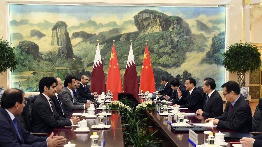 L'image montre une réunion formelle dans une salle de conférence. On peut y voir plusieurs personnes assises autour d'une longue table. À l'arrière-plan, il y a deux drapeaux, celui du Qatar et celui de la Chine, ainsi qu'une grande fresque représentant des montagnes et des paysages. Les participants semblent engagés dans une discussion, avec des tasses et des documents disposés sur la table. L'ambiance semble sérieuse et professionnelle, indiquant un dialogue diplomatique entre les deux pays.