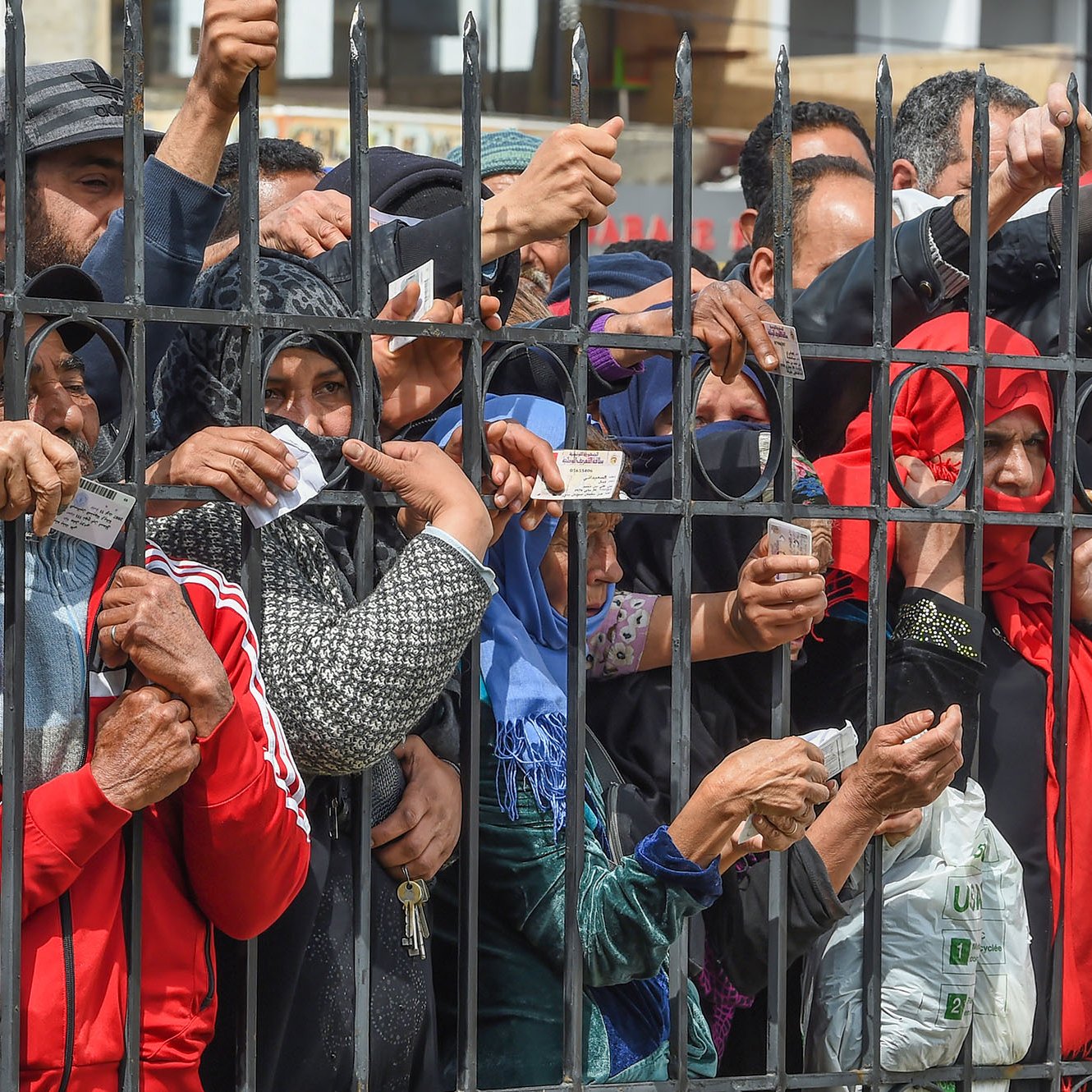 L'image montre un groupe de personnes rassemblées derrière des barreaux, semblant impatientes ou en attente. On peut voir des mains tendues à travers les barreaux, certains tenant des objets comme des cartes ou des papiers. Les visages de quelques personnes sont également visibles, exprimant diverses émotions. L'ambiance semble tendue, suggérant une situation de demande ou d'attente d'assistance. Les vêtements des individus montrent une diversité de styles, et il y a une impression de solennité dans la scène.