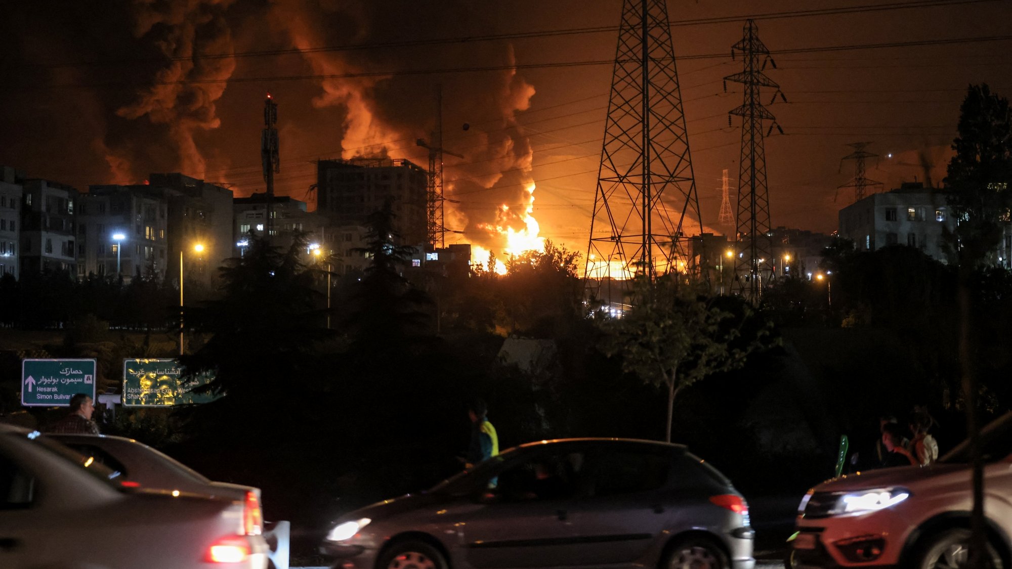 Un forte incendio illumina il cielo notturno, con fumi neri e auto in movimento.