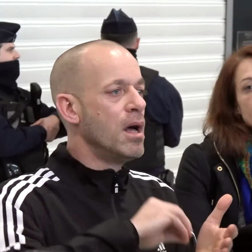 L'image montre un homme qui parle, visiblement en train d'expliquer quelque chose avec des gestes. Il porte un survêtement noir avec des rayures blanches. À côté de lui, une femme l'écoute attentivement, les cheveux bruns et portant un manteau noir. À l'arrière-plan, on peut voir des policiers en uniforme, certains portant des masques. L'ensemble suggère une situation de discussion ou d'intervention, possiblement liée à un événement impliquant les forces de l'ordre.