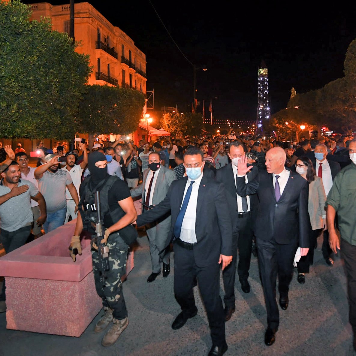 تظهر الصورة مجموعة من الأشخاص في شوارع مدينة مضاءة بأضواء ليلية. يتواجد في المقدمة رجل يرتدي بدلة مع أشخاص آخرين، من بينهم رجل ملثم يحمل سلاحًا. يبدو أن هناك تجمعًا من الحشود في الخلفية، حيث يتفاعل بعضهم مع الموقف. الأجواء توحي بوجود حدث أو مظاهرة، ومشاعر الحماس والانفعال واضحة في تعابير الحضور.