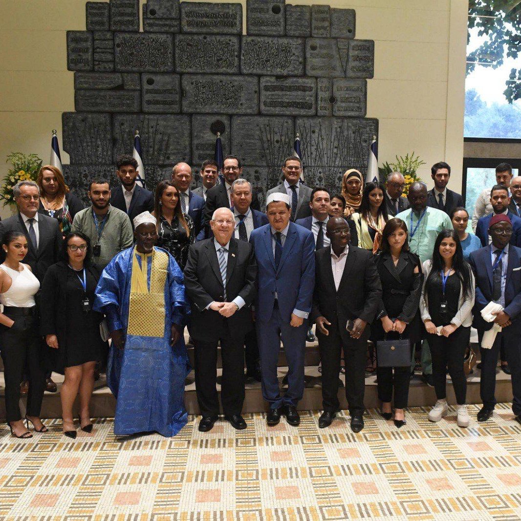 The image shows a large group of people gathered together for a formal event. They are standing in a room with a decorative backdrop that includes flags and a large, textured wall. The group consists of men and women of diverse backgrounds, and many are dressed in professional attire. Some individuals wear traditional cultural clothing. Everyone appears to be smiling, and they are posed together for a photograph, likely to commemorate the occasion. The setting is bright and well-lit, contributing to a positive atmosphere.