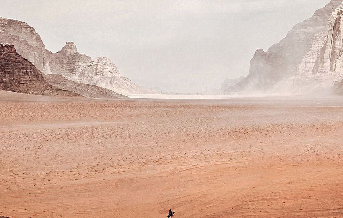 L'image montre un vaste paysage désertique. On peut voir des montagnes rocheuses au loin, entourées de dunes de sable. Le ciel est nuageux, ce qui crée une atmosphère légèrement brumeuse. Au centre, une seule silhouette humaine, probablement un cavalier, se déplace à travers ce paysage aride. La scène transmet une sensation d'immensité et de solitude.