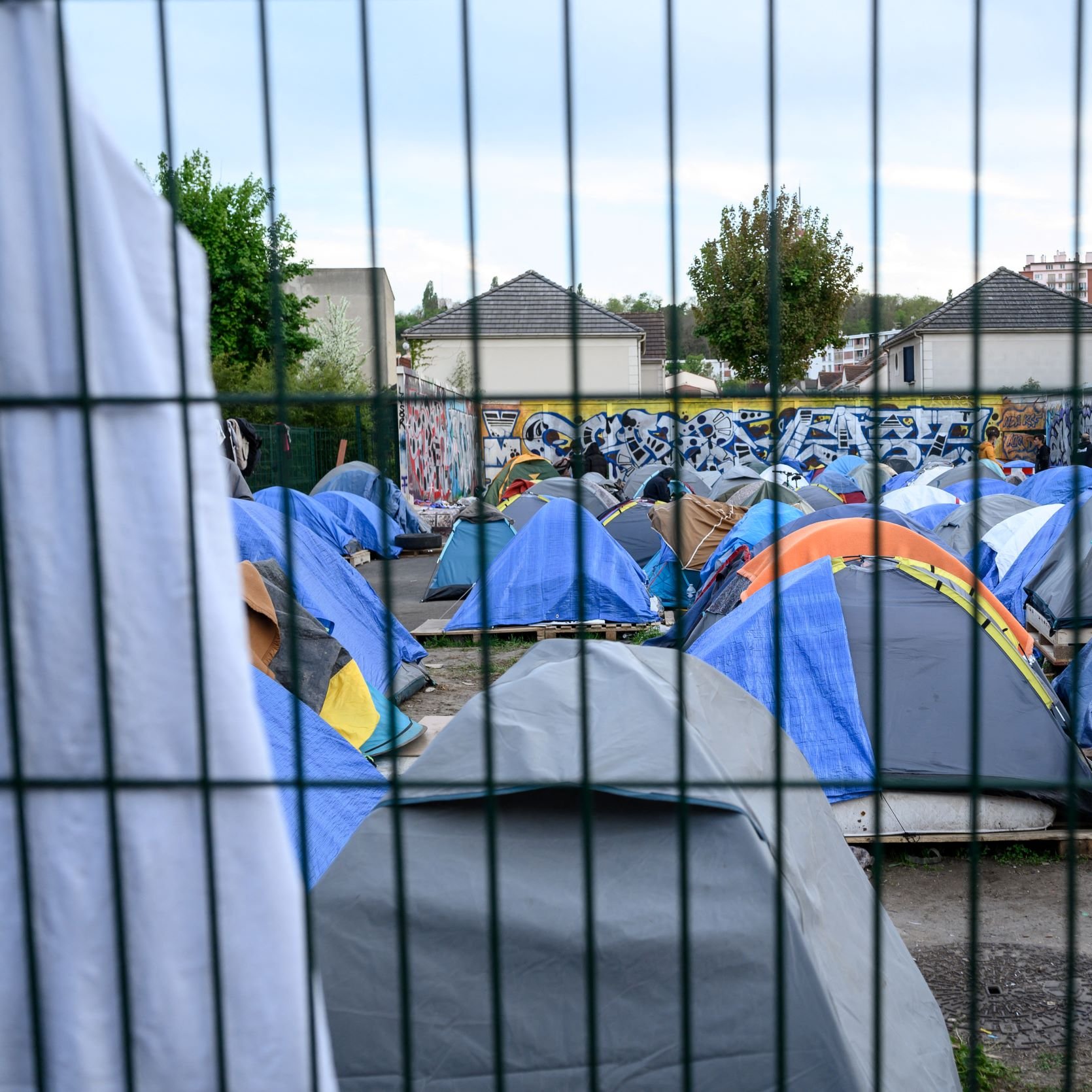 این تصویر نمایی از یک اردوگاه چادرها را نشان می‌دهد. چادرهای متعدد با رنگ‌های مختلف در یک فضای باز قرار دارند و برخی از افراد در اطراف آن‌ها مشغول فعالیت هستند. در پس‌زمینه، یک دیوار با هنر خیابانی یا گرافیتی قابل مشاهده است. همچنین، یک نرده فلزی در جلوی تصویر وجود دارد که فضا را از محیط بیرونی جدا می‌کند. هوای آسمان می‌تواند کمی ابری به نظر برسد.