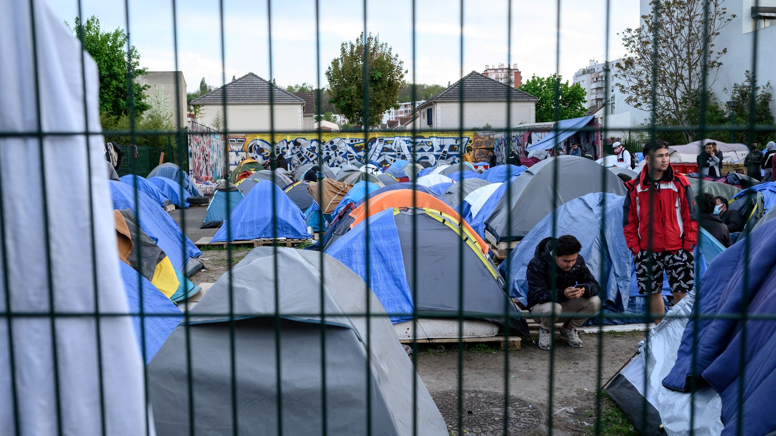 L'image montre un campement de tentes installé dans une zone urbaine. Les tentes sont de différentes tailles et couleurs, principalement en bleu et gris. On peut voir des personnes présentes, certaines debout et d'autres assises, semblant utiliser leur téléphone. En arrière-plan, il y a des bâtiments et un mur décoré de graffitis. L'ensemble du camp est entouré par une clôture, ce qui donne un aspect de confinement à la scène. Les arbres et la végétation ajoutent une touche de verdure au décor.