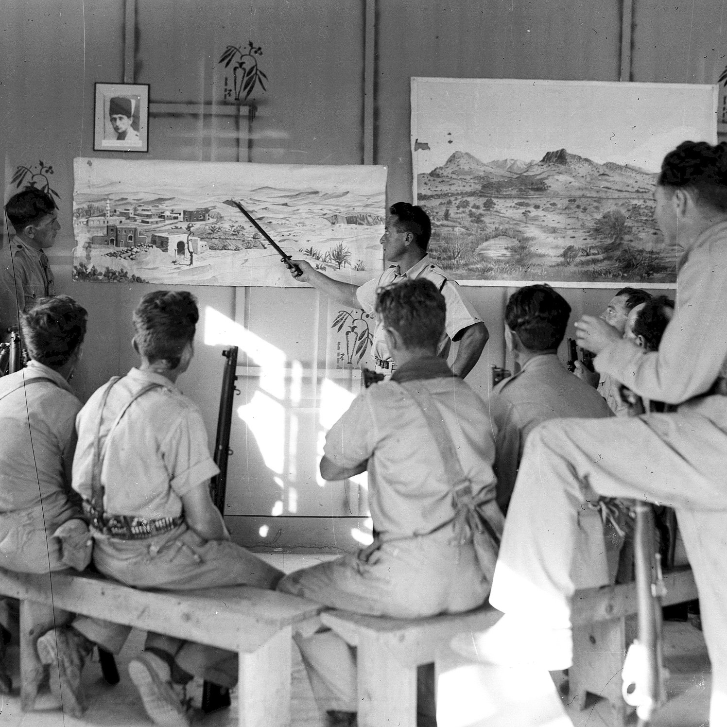 L'image montre un groupe de personnes assises sur des bancs, attentives à un homme qui semble donner une présentation devant des tableaux accrochés au mur. Les tableaux illustrent des paysages, l'un représentant probablement un village ou une scène de la nature. L'environnement est dépouillé, avec des murs simples et une lumière naturelle qui pénètre à travers une fenêtre. Les participants, majoritairement en vêtements uniformes, semblent captivés par l'explication de l'intervenant. Il y a aussi un portrait encadré sur le mur, ajoutant un élément de contexte à la scène.