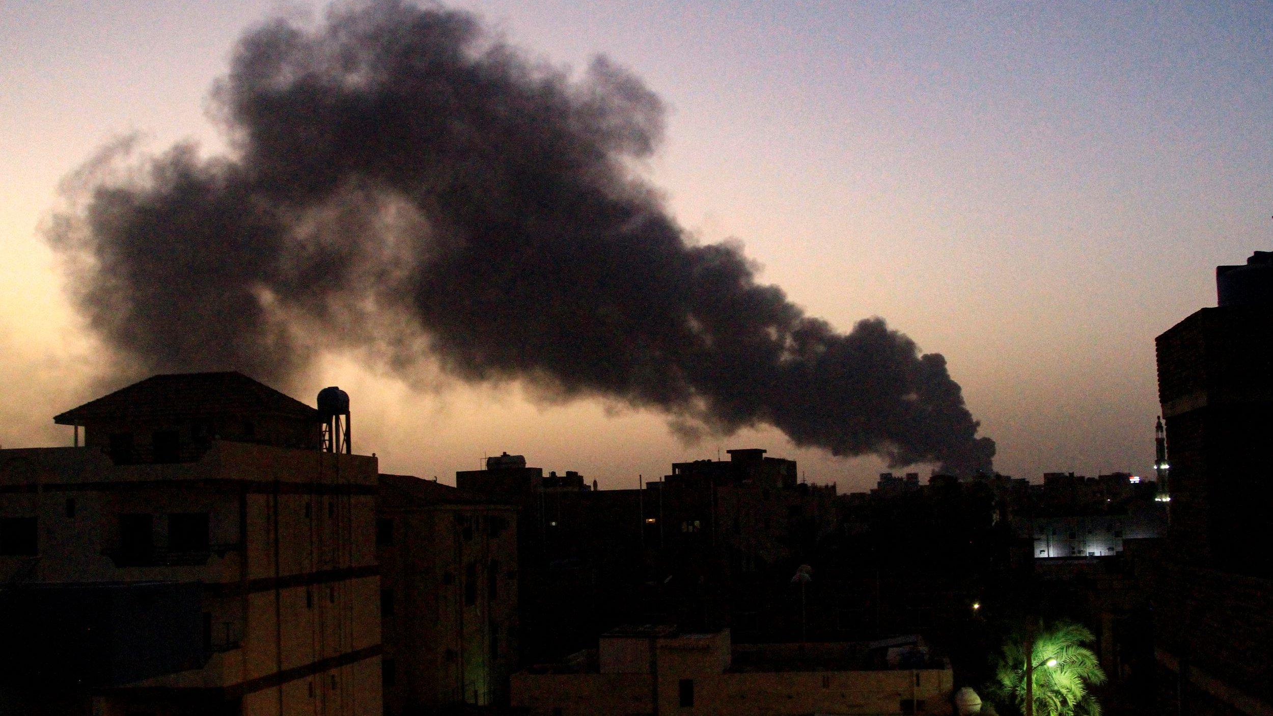 The image depicts a dusk or evening scene in an urban environment, with several buildings visible in the foreground. In the background, a substantial plume of black smoke rises into the sky, creating a stark contrast against the fading light of sunset. The smoke suggests a fire or some kind of industrial activity. The overall mood of the image conveys a sense of urgency or concern, given the presence of the smoke.