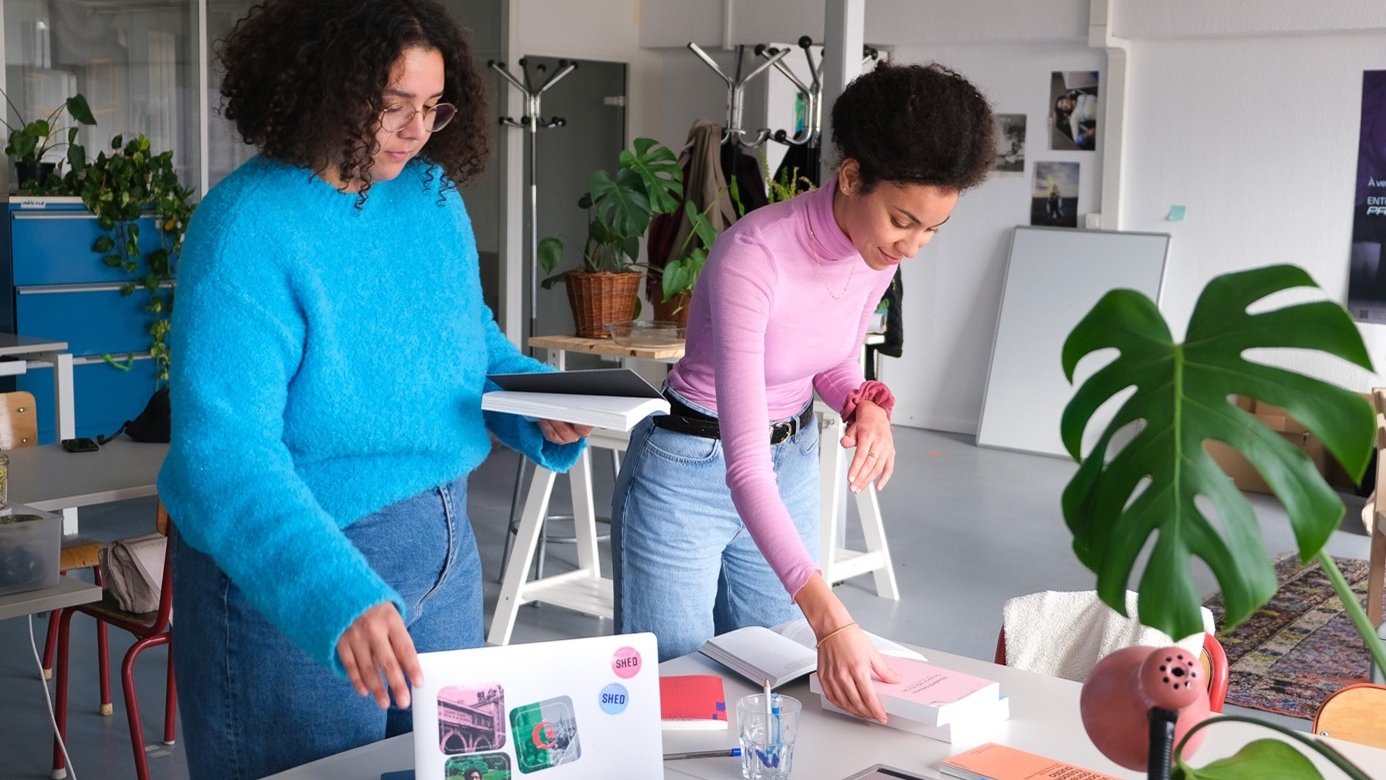 L'image montre deux femmes dans un environnement de travail créatif. L'une d'elles, vêtue d'un pull bleu, semble arranger des choses sur une table. L'autre femme, en pull rose, est en train de manipuler des livres ou des documents. Sur la table, il y a un ordinateur portable, des livres et quelques accessoires de bureau. L'espace est lumineux et décoré de plantes, ce qui contribue à une ambiance agréable et inspirante.