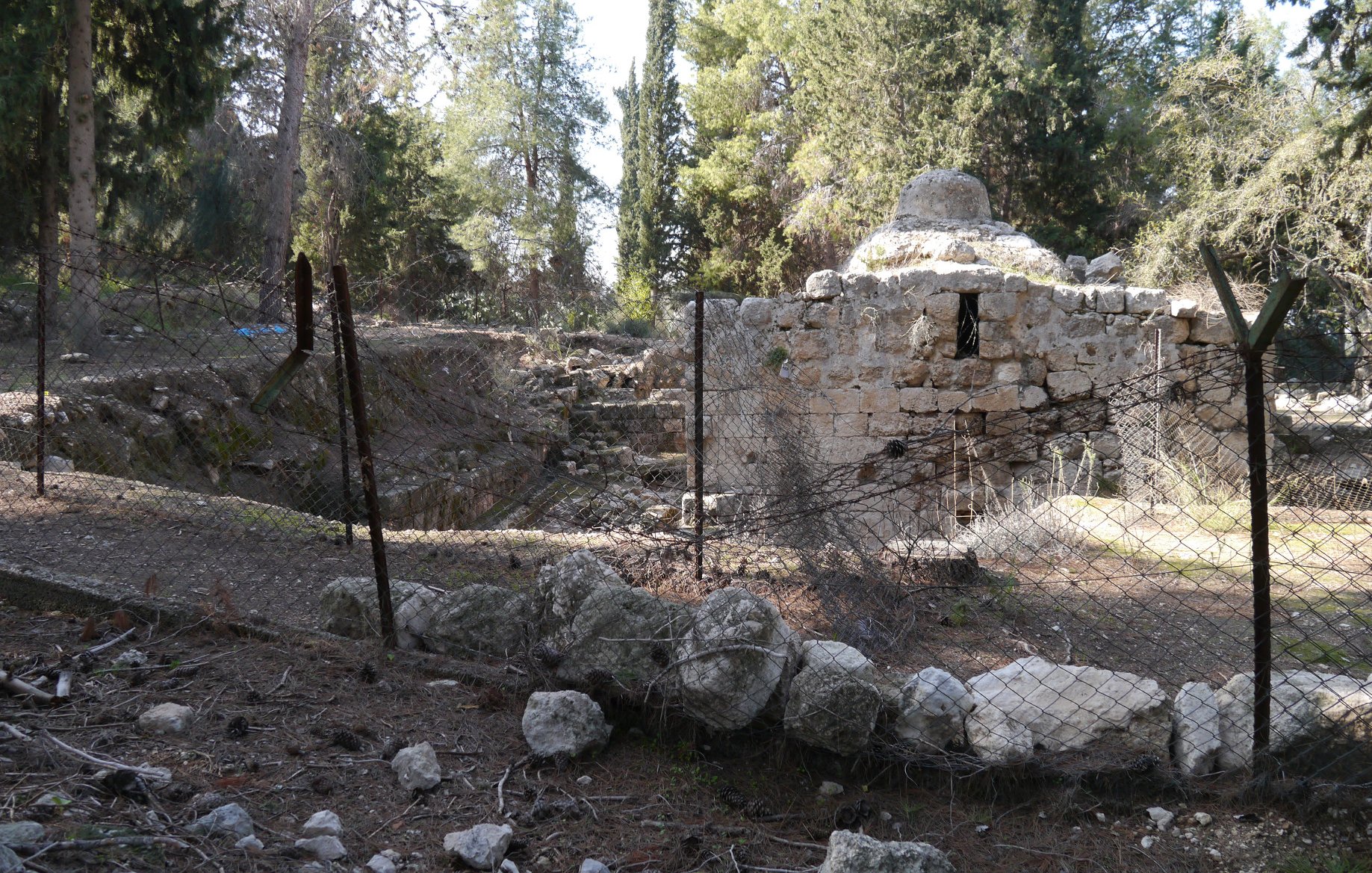 L'image montre un site archéologique entouré d'une clôture en fil de fer. Il y a des ruines anciennes, probablement d'une structure en pierre, avec des éléments végétaux environnants tels que des arbres et des buissons. Le sol est rempli de pierres et de débris, ce qui donne une impression d'abandon. L'atmosphère semble paisible, mais aussi légèrement mélancolique, témoignant d'un passé oublié.