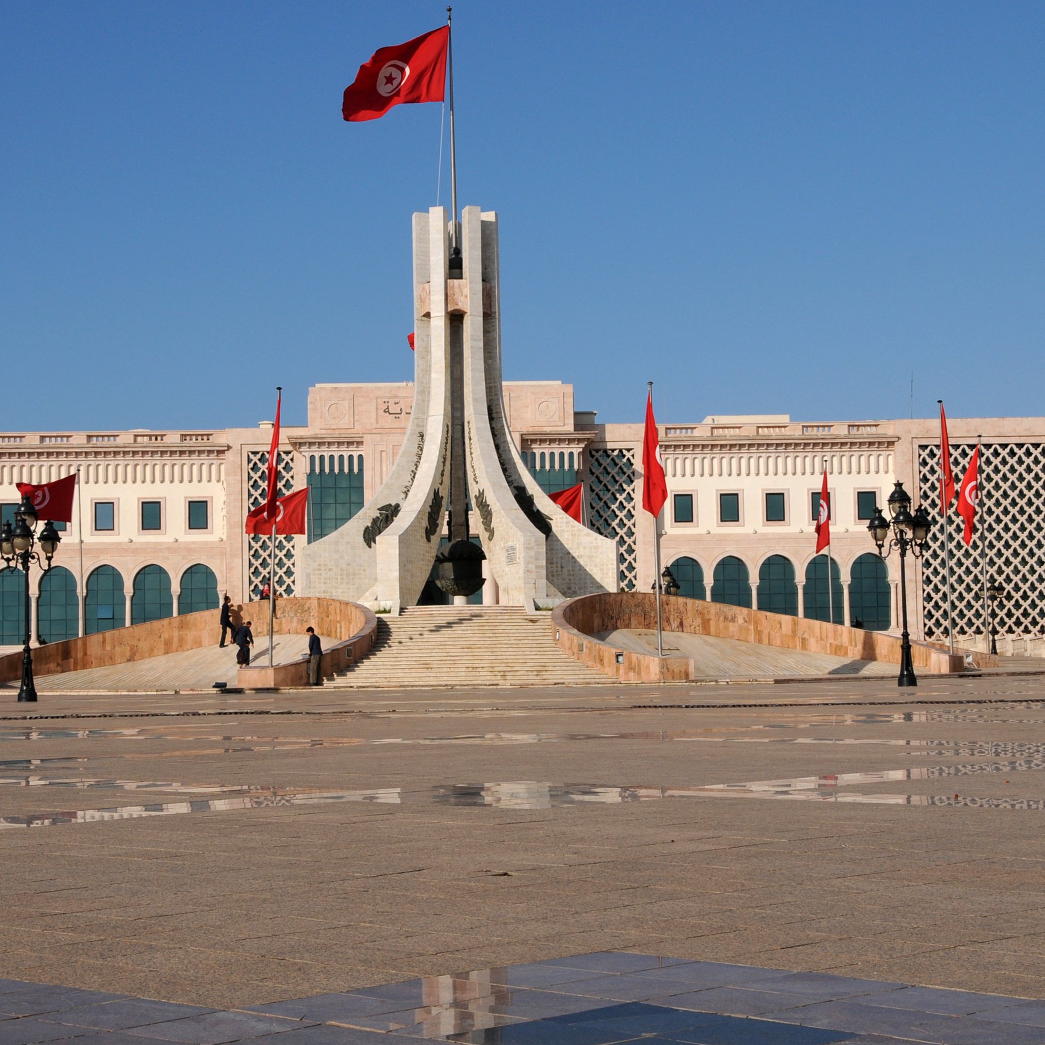 L'image montre un bâtiment imposant avec une architecture distinctive, situé probablement dans un espace public. Au centre, on peut voir un mât portant le drapeau tunisien, avec plusieurs drapeaux rouges autour. L'esplanade devant le bâtiment est pavée, et des lampadaires élégants sont présents sur les côtés. Le ciel est dégagé, ce qui donne une atmosphère lumineuse et claire à la scène. L'architecture semble moderne mais avec des éléments traditionnels.