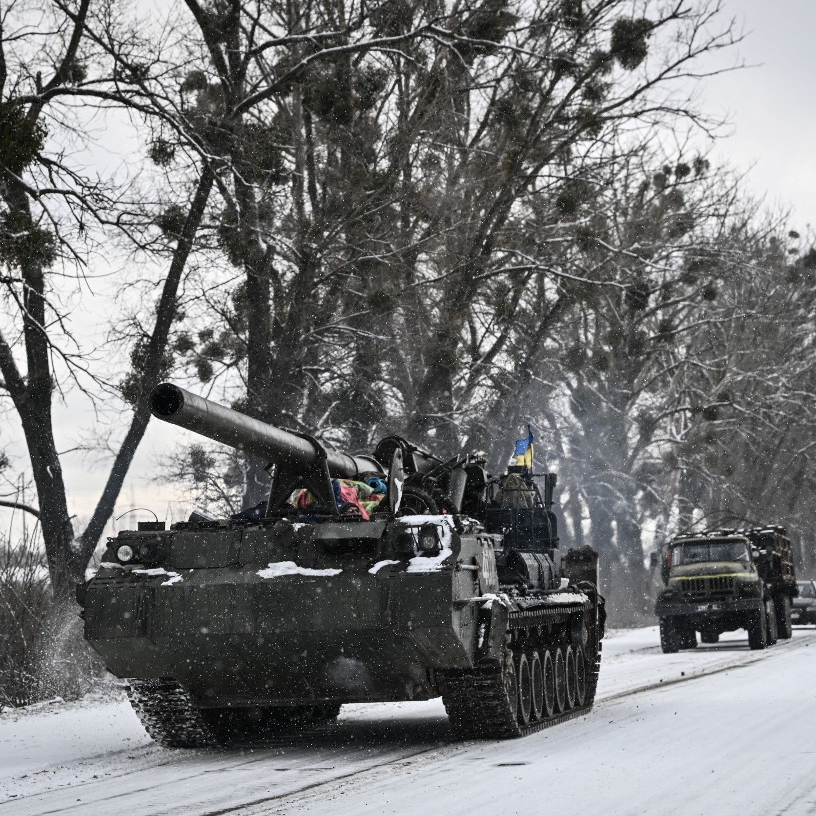 L'image montre un paysage hivernal avec de la neige recouvrant le sol. Un char militaire, probablement un tank, avance sur une route étroite bordée d'arbres. Des véhicules militaires sont également visibles en arrière-plan, créant une impression de mouvement dans un environnement froid et désert. L'atmosphère est marquée par des nuances de gris et de blanc, typiques d'un paysage d'hiver.