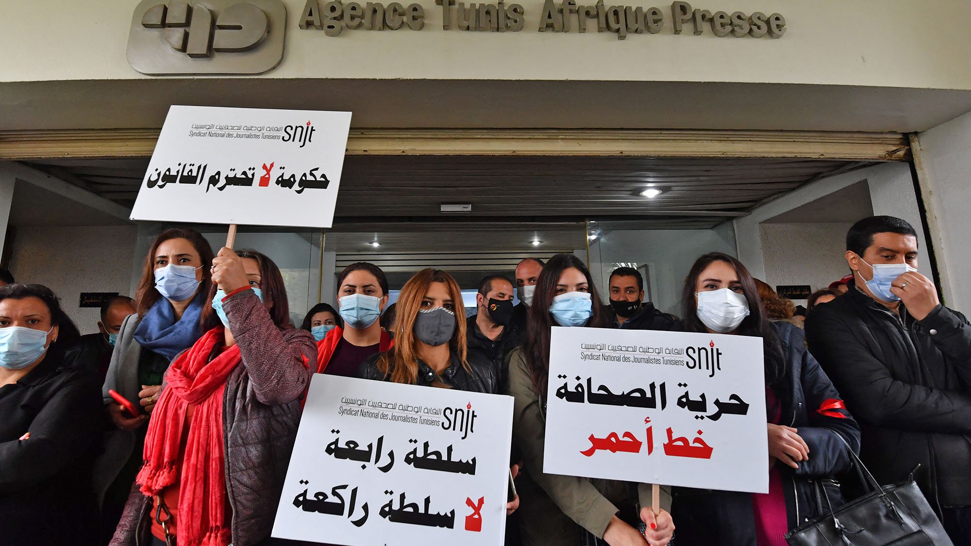 L'image montre un groupe de femmes et d'hommes rassemblés devant un bâtiment, portant des pancartes. Les pancartes expriment des messages liés à la liberté de la presse et critiquent le gouvernement. Les manifestants portent des masques, probablement en raison de préoccupations sanitaires. En arrière-plan, on peut voir le logo d'une agence de presse. L'atmosphère semble déterminée, avec un engagement visible pour des causes liées à la liberté d'expression.