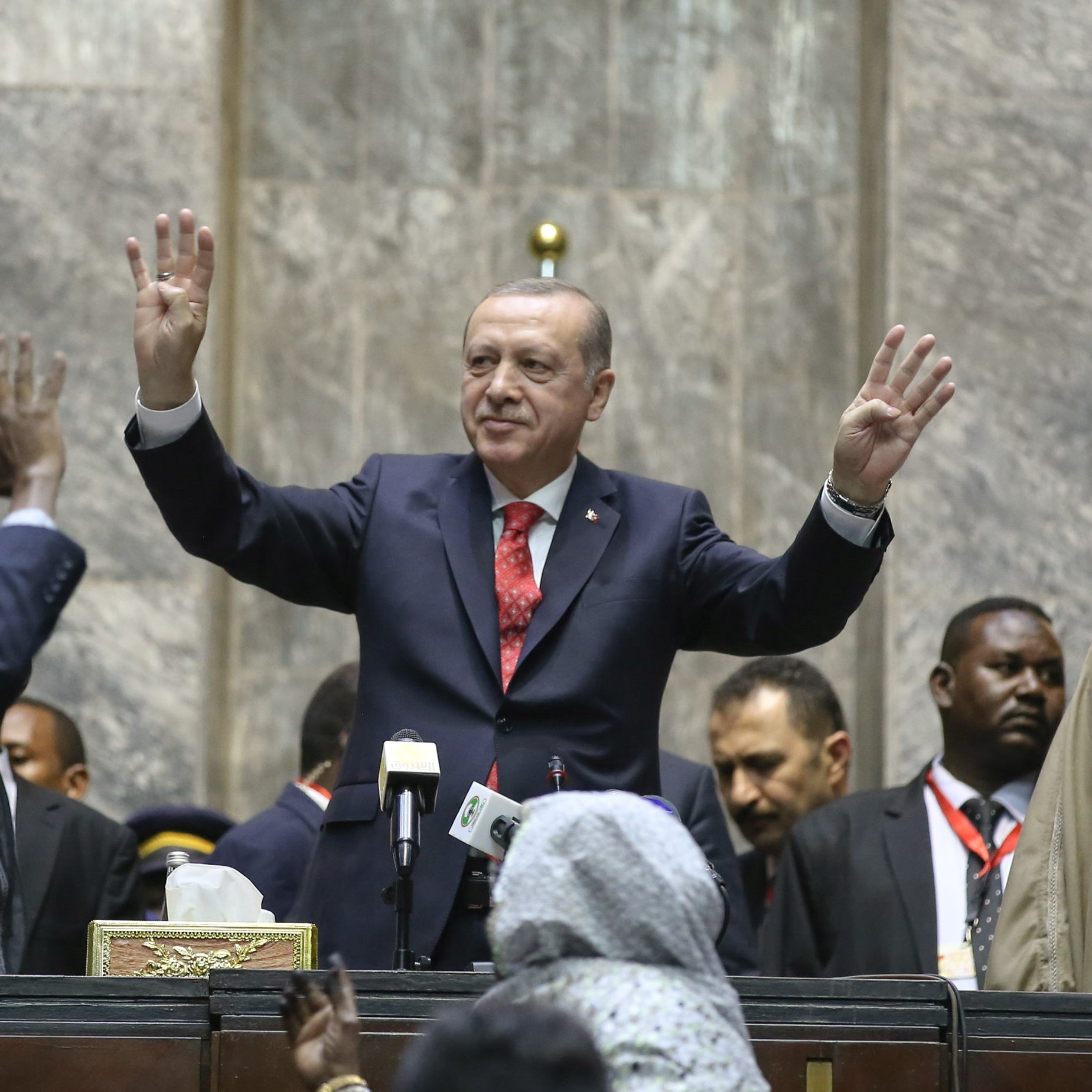 تظهر الصورة تجمعًا سياسيًا حيث يقف شخصية معروفة في المنتصف، مرتديًا بدلة رسمية، ويقوم بإشارة بيديه. خلفه يظهر عدد من الأشخاص، بعضهم يبدو متحمسًا ويرفع أيديه، بينما يوجد آخرون يقفون بجانبه. المكان يبدو رسميًا، مع وجود خلفية مكونة من جدران حجرية وأعلام. الأجواء تعكس حماس الحضور.