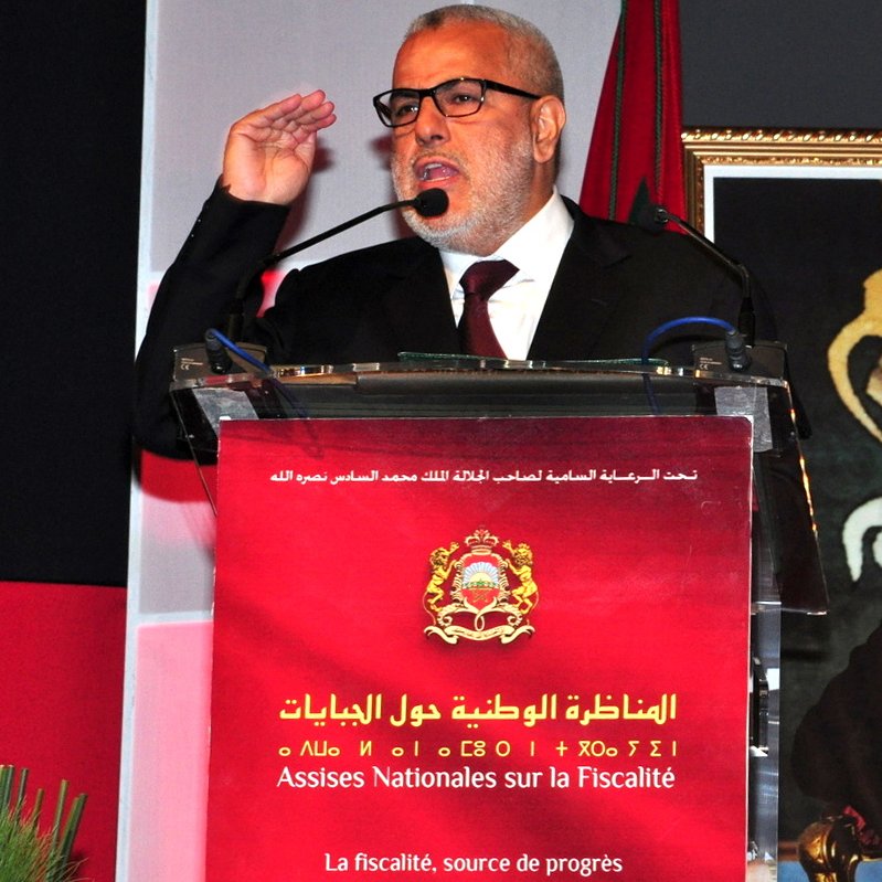 الصورة تظهر شخصًا معروفًا يتحدث على منصة، حيث يبدو أنه يلقي خطابًا رسميًا. خلفه يمكن رؤية شعار رسمي أو صورة رمزية. المنصة تحمل عنوان "المناظرة الوطنية حول الأداءات"، مما يشير إلى أن الحدث يتعلق بالمالية أو الضرائب. الجو العام يبدو رسميًا وجديًا، ولغة الجسد تعكس الاهتمام والالتزام.