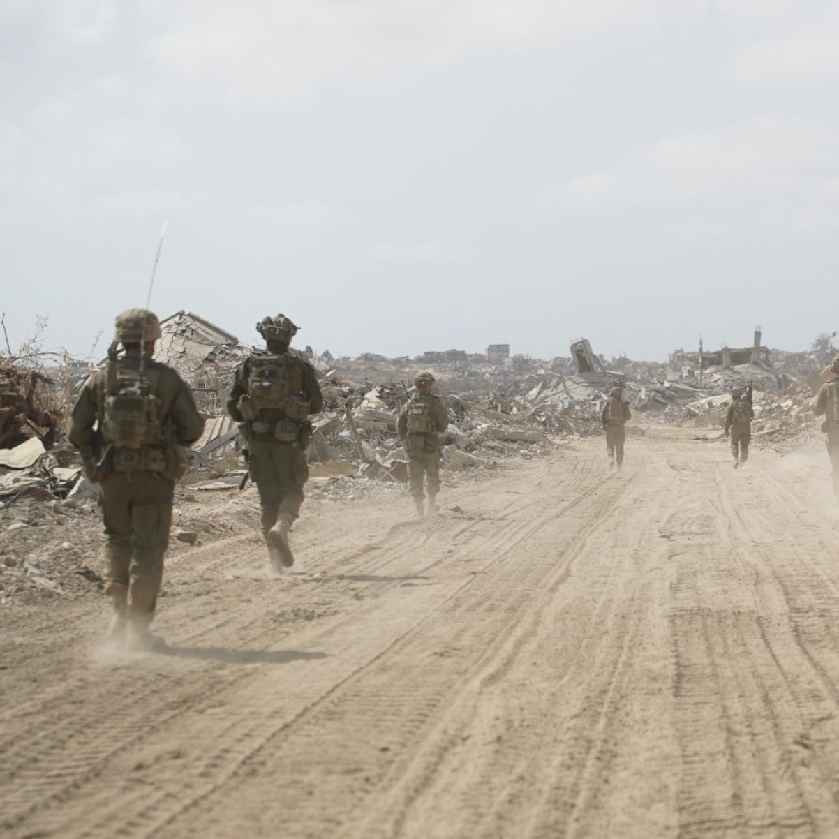 Soldiers in military gear walk through a dusty, desolate landscape with debris around them.