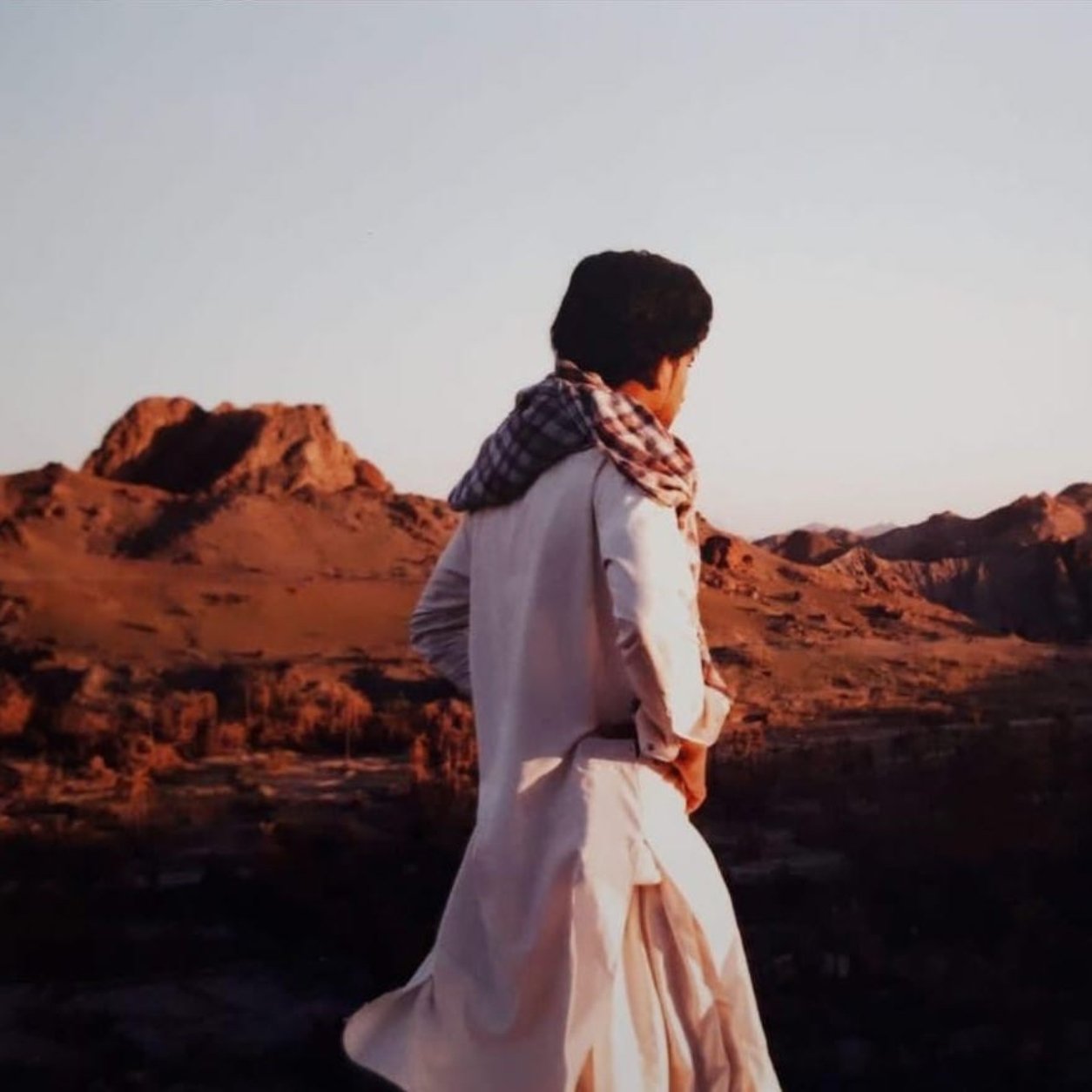 در این تصویر، فردی با لباس سنتی و شال بر دوش، به سمت یک چشم‌انداز طبیعی نگاه می‌کند. زمینه تصویر شامل کوه‌های بلند و دشت‌های وسیع است که نور ملایم غروب خورشید بر آنها تابیده شده است. رنگ‌های گرم غروب و سایه‌های بلند کوه‌ها زیبایی خاصی به صحنه داده‌اند. احساس آرامش و سکوت در فضای تصویر به وضوح حس می‌شود.