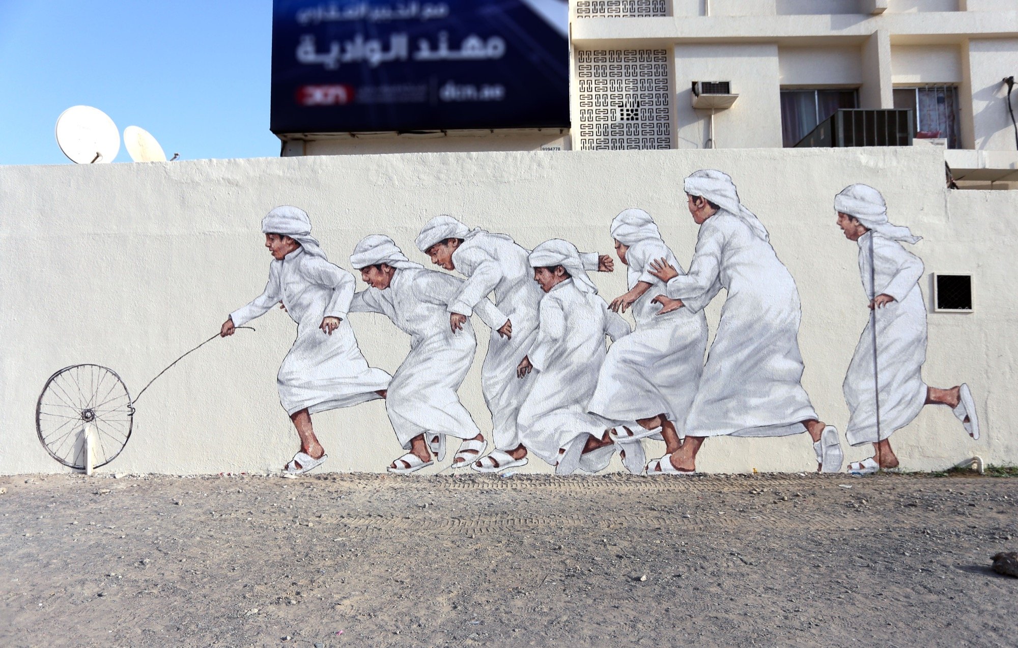 L'image montre une fresque murale représentant plusieurs enfants en vêtements traditionnels blancs. Ils semblent courir joyeusement, certaines figures tenant un fil ou une roue, ce qui évoque un jeu. Le décor est d'un style urbain, avec un fond minimaliste et une ambiance dynamique. L'art mural met en valeur la culture et la joie de l'enfance.