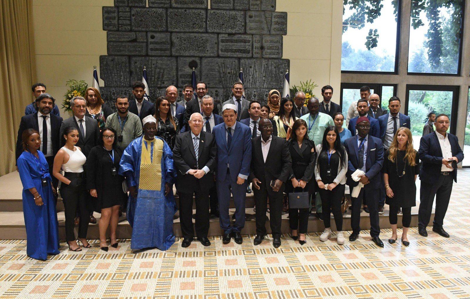 L'image montre un groupe de personnes réunies, probablement pour un événement officiel ou une cérémonie. Au premier plan, on voit des hommes en costume et quelques femmes en tenues formelles. Certains portent des vêtements traditionnels, ce qui suggère une diversité culturelle parmi les participants. En arrière-plan, on remarque des drapeaux et une décoration murale. L'ambiance semble conviviale et professionnelle, avec des sourires et des échanges amables entre les membres du groupe.