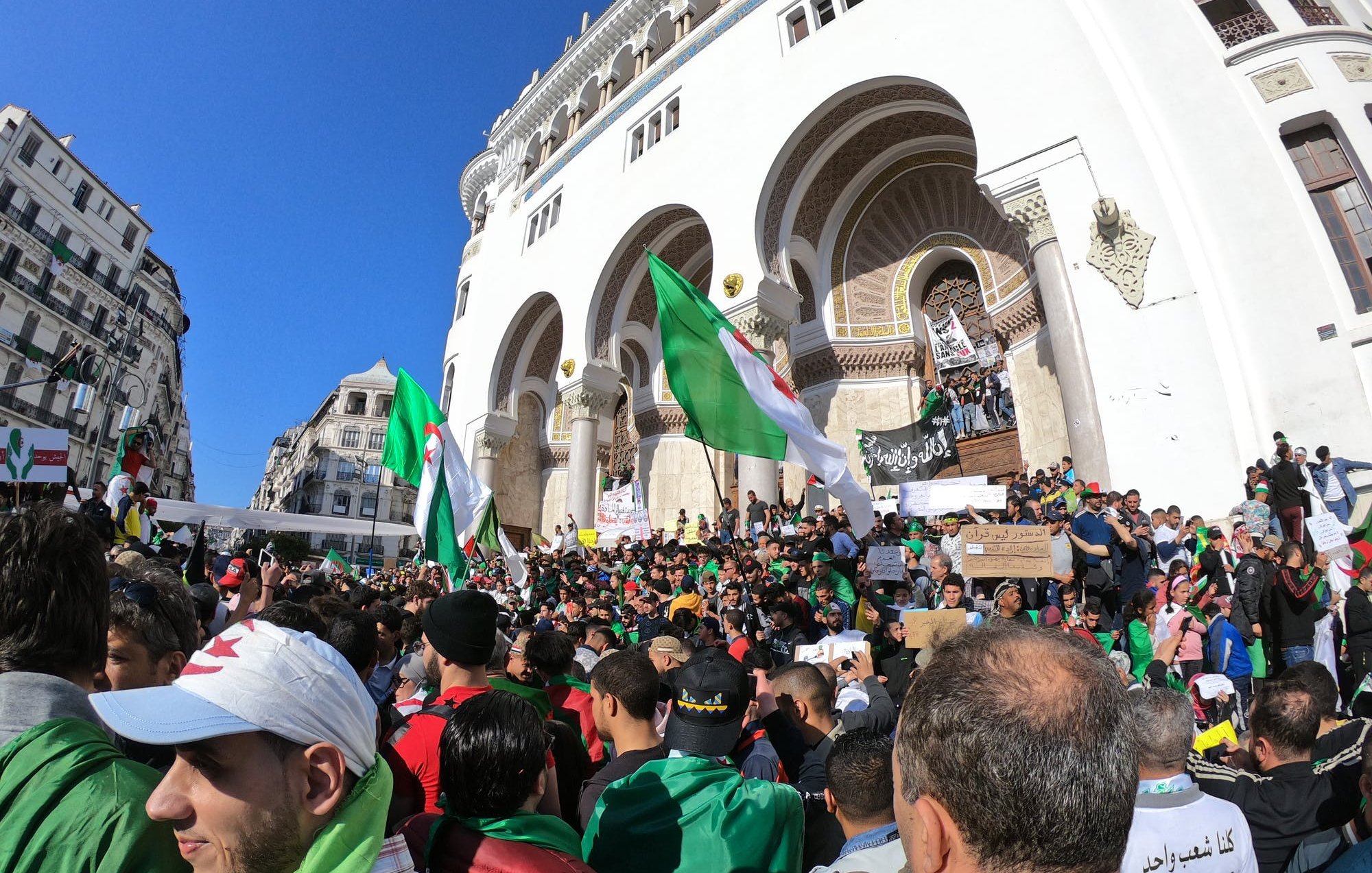 L'image montre une grande foule rassemblée devant un bâtiment avec une architecture traditionnelle. Les manifestants brandissent des drapeaux algériens et des pancartes, exprimant probablement des revendications politiques ou sociales. La scène est en plein air, sous un ciel bleu, et il y a une atmosphère de mobilisation collective, avec des personnes de tous âges vertes et rouges.