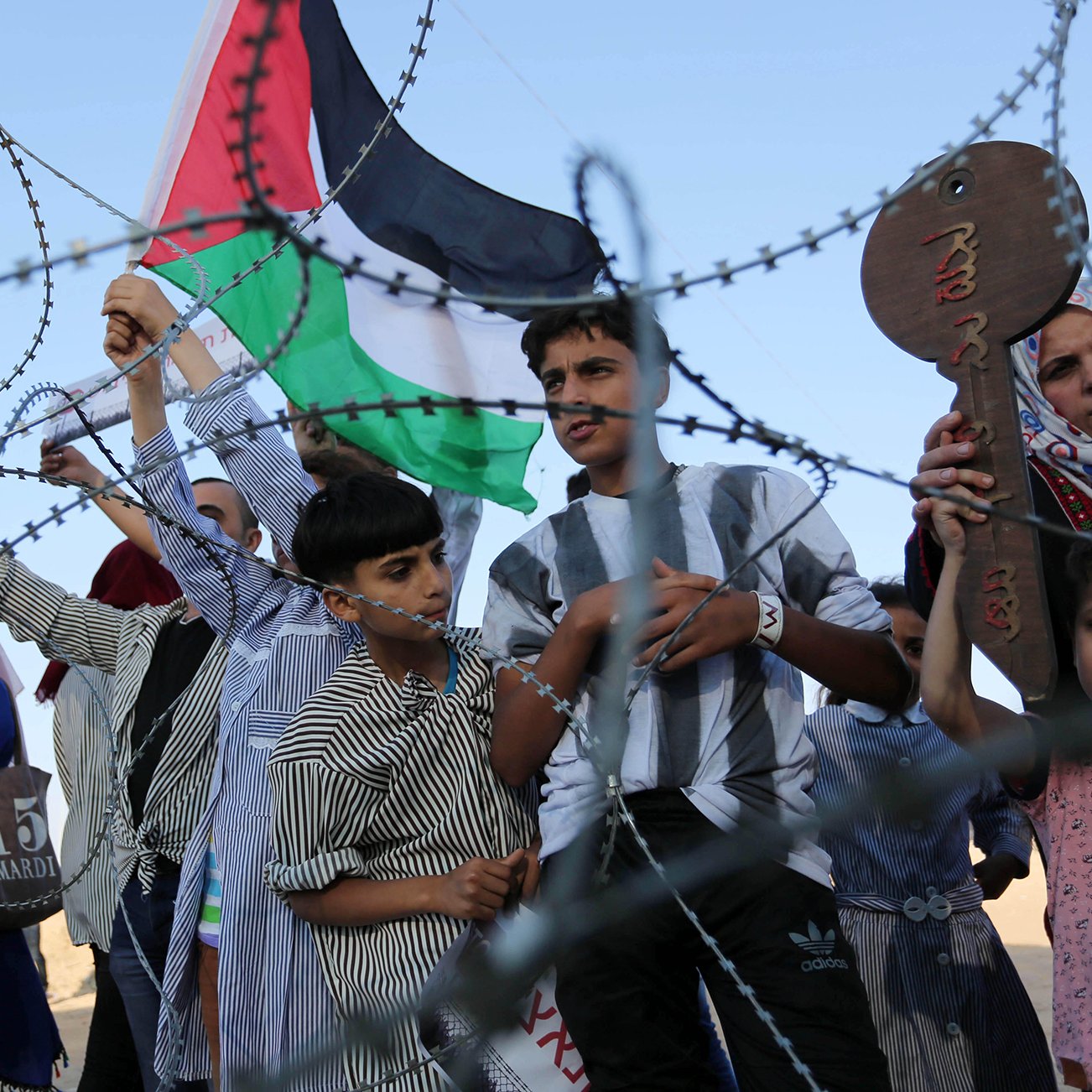 L'image montre un groupe de personnes, y compris des enfants, qui tiennent des drapeaux palestiniens et des pancartes. Ils se tiennent derrière une barrière semblable à du barbelé. Les expressions des personnes peuvent communiquer des émotions telles que la détermination ou l'espoir. L'environnement semble être en plein air, sans autre détail contextuel visible.