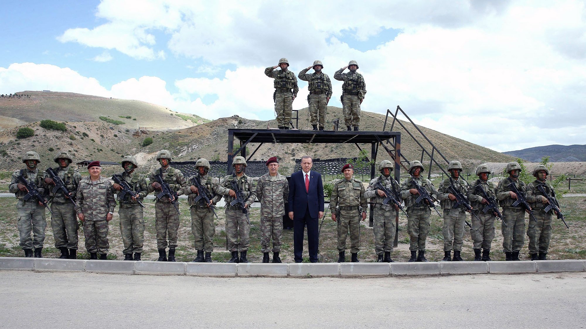 La imagen muestra a un grupo de soldados en un entorno militar. Hay un total de quince soldados alineados, vestindo uniformes de camuflaje y sosteniendo sus armas. En el centro de la imagen, se encuentra un hombre vestido con traje, aparentemente en una posición de liderazgo. Al fondo, se pueden ver dos soldados más en una plataforma elevada, saludando en posición de firme. El paisaje que rodea al grupo es montañoso y despejado, con nubes en el cielo.