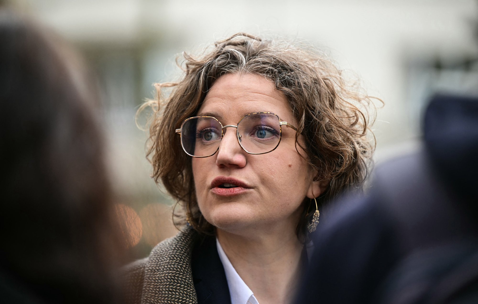 L'image montre une femme avec des cheveux bouclés et des lunettes, en train de parler. Elle semble engagée dans une conversation, peut-être lors d'un événement public. Son expression faciale est sérieuse, et elle porte un vêtement avec un col blanc et une veste. L'arrière-plan paraît flou, suggérant qu'elle se trouve au milieu d'un groupe ou d'une foule.