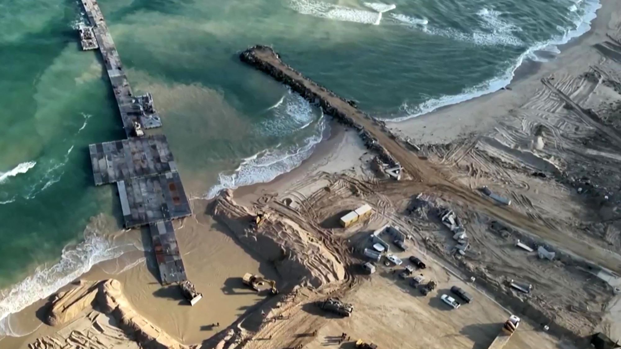 L'immagine mostra una vista aerea di una zona costiera in fase di costruzione o ristrutturazione. Si possono vedere macchine da lavoro e attrezzature presenti sulla spiaggia e nelle vicinanze, mentre un molo si estende verso il mare. Le onde del mare si infrangono sulla riva, e il terreno circostante appare movimentato e scavato, con evidenti segni di lavori in corso. La luce naturale esalta i dettagli del paesaggio, creando un contrasto tra l'acqua blu e la sabbia.